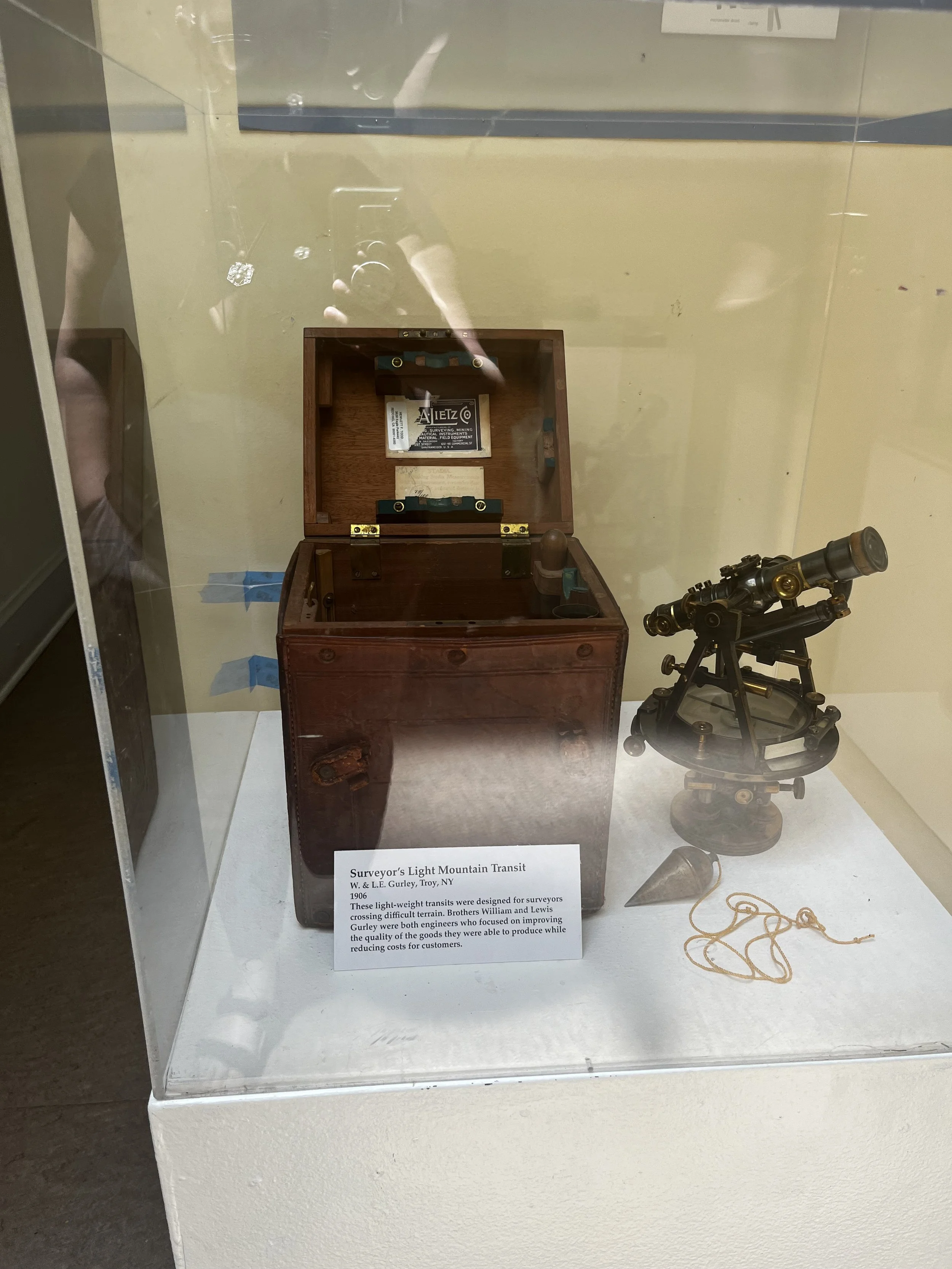 A display case containing a vintage surveyor's light mountain transit, a wooden box, and a small metallic telescope with a rope tied to it. There is a small informational placard describing the objects.
