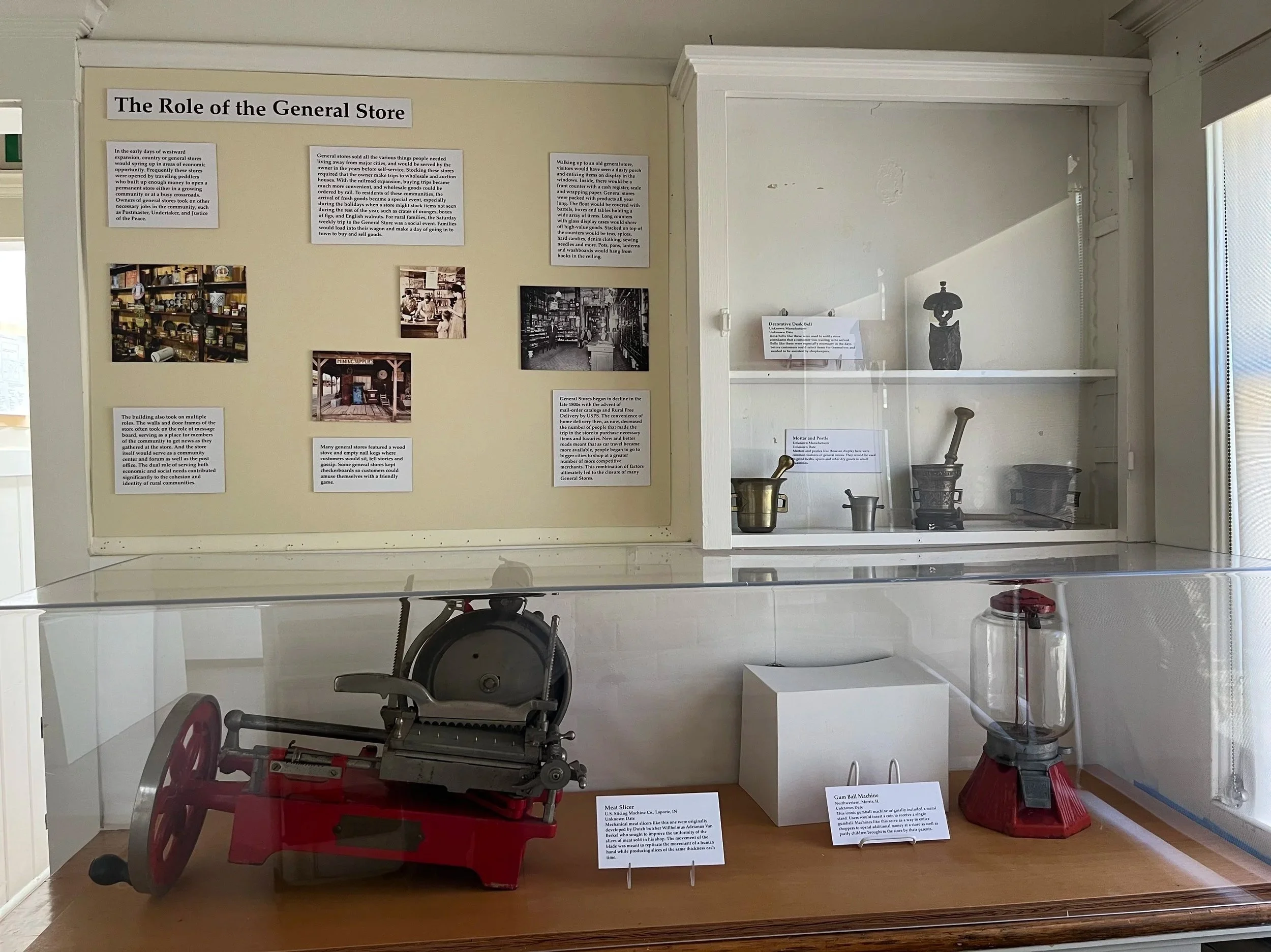 Exhibit of vintage kitchen tools including a meat slicer, gum ball machine, and a jar, displayed behind a glass case with informational placards.
