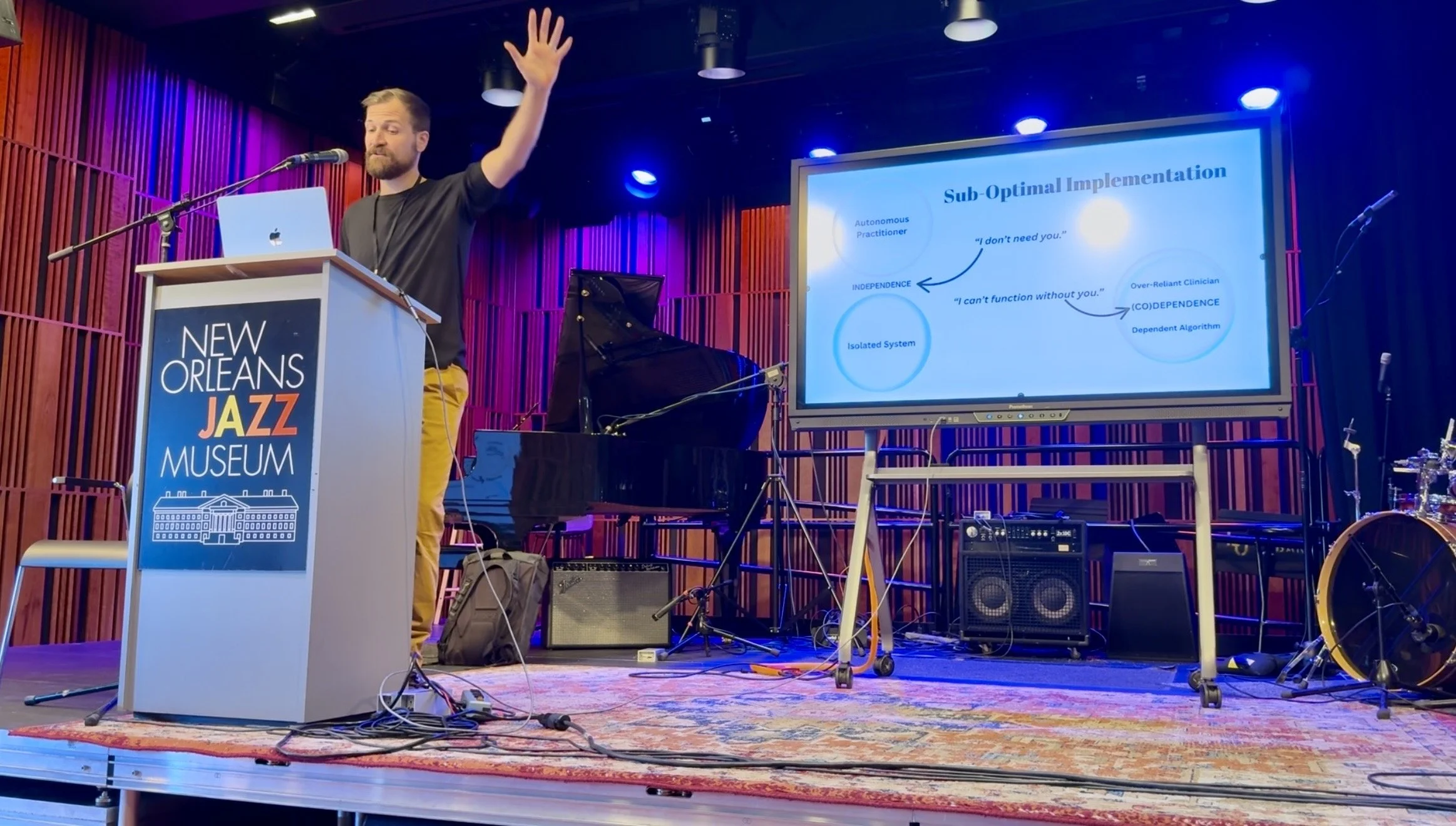 A man giving a presentation at the New Orleans Jazz Museum with a podium, a large screen displaying a diagram, and musical instruments on stage.
