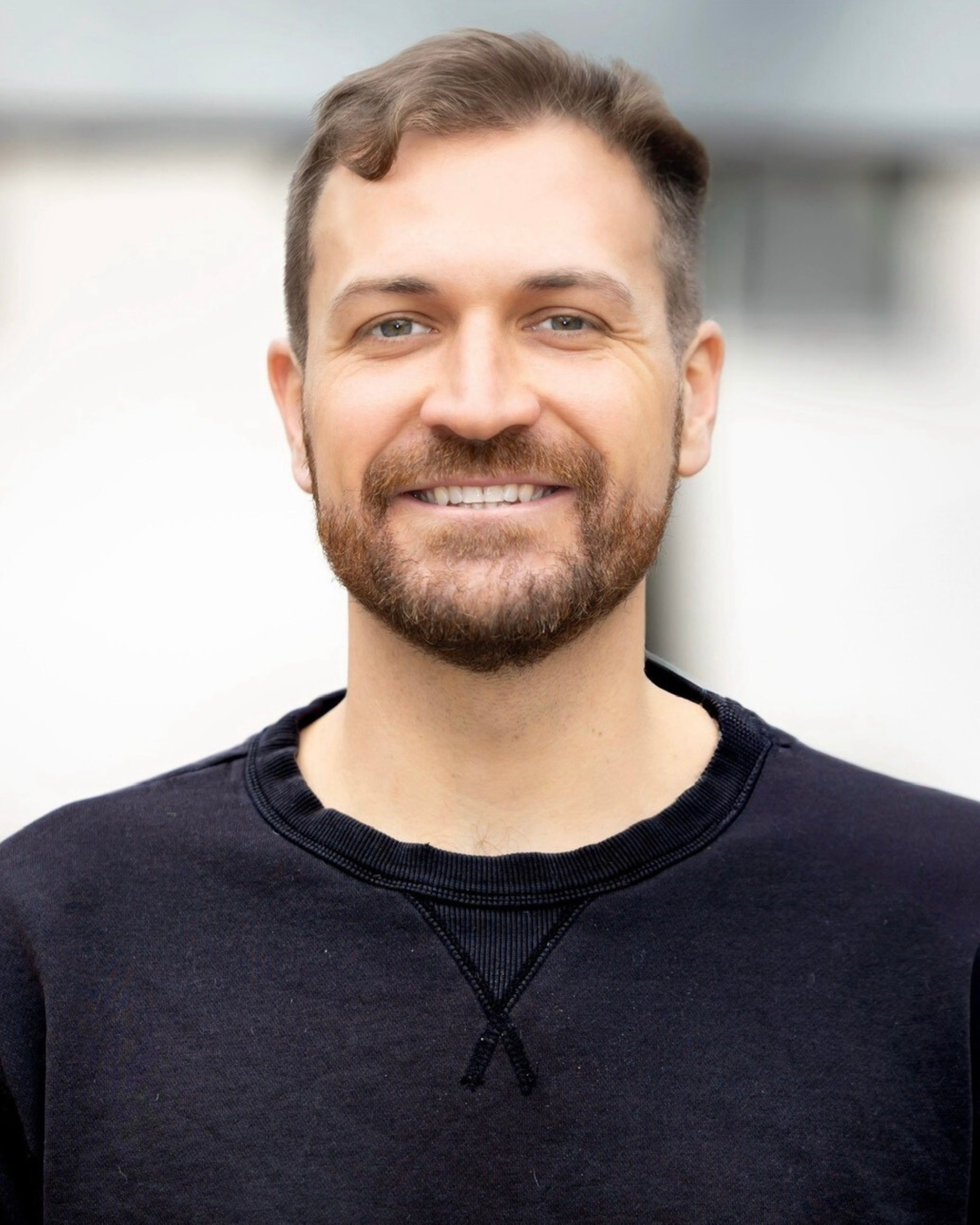 A smiling man with a beard and short brown hair, wearing a black crew neck sweatshirt, standing outdoors with a blurred background.