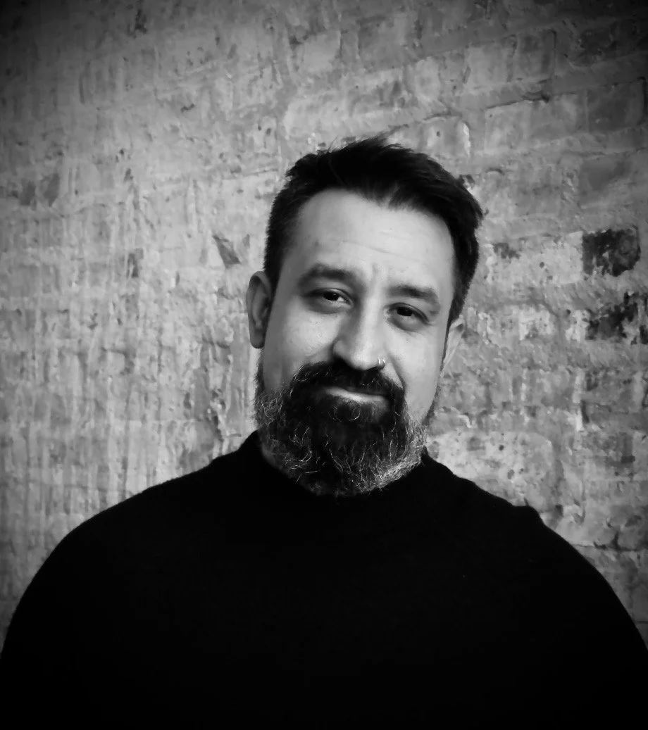 Black and white portrait of a man with a beard, mustache, and nose ring, wearing a dark turtleneck sweater, standing in front of a textured brick wall.