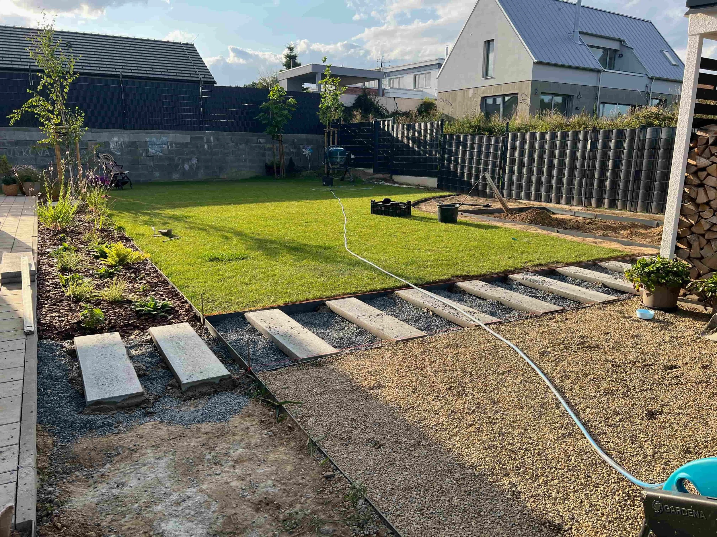 Rozpracovaná zahrada při realizaci – trávník, květinové záhony, dlažba a stavební materiál u moderního domu.