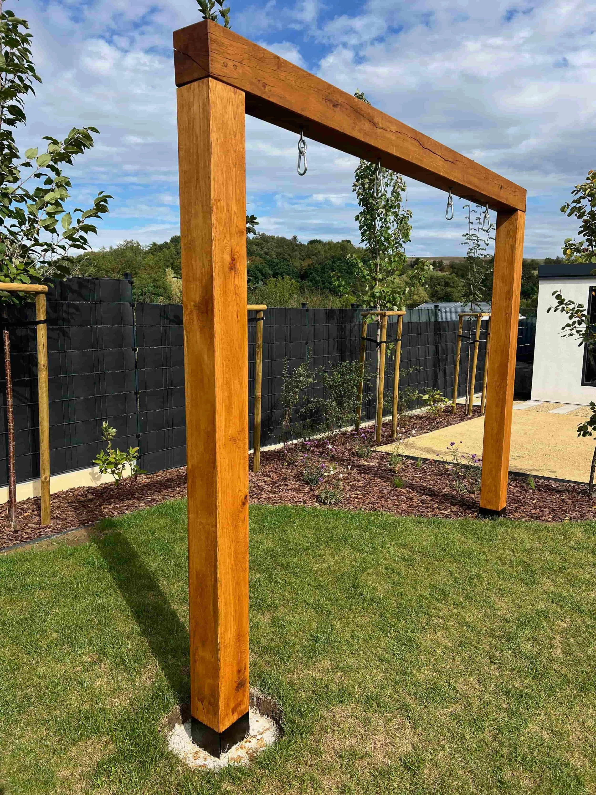 Dřevěná venkovní konstrukce pro cvičení s kovovými závěsnými prvky, umístěná na zahradě s trávníkem a mladými stromy.