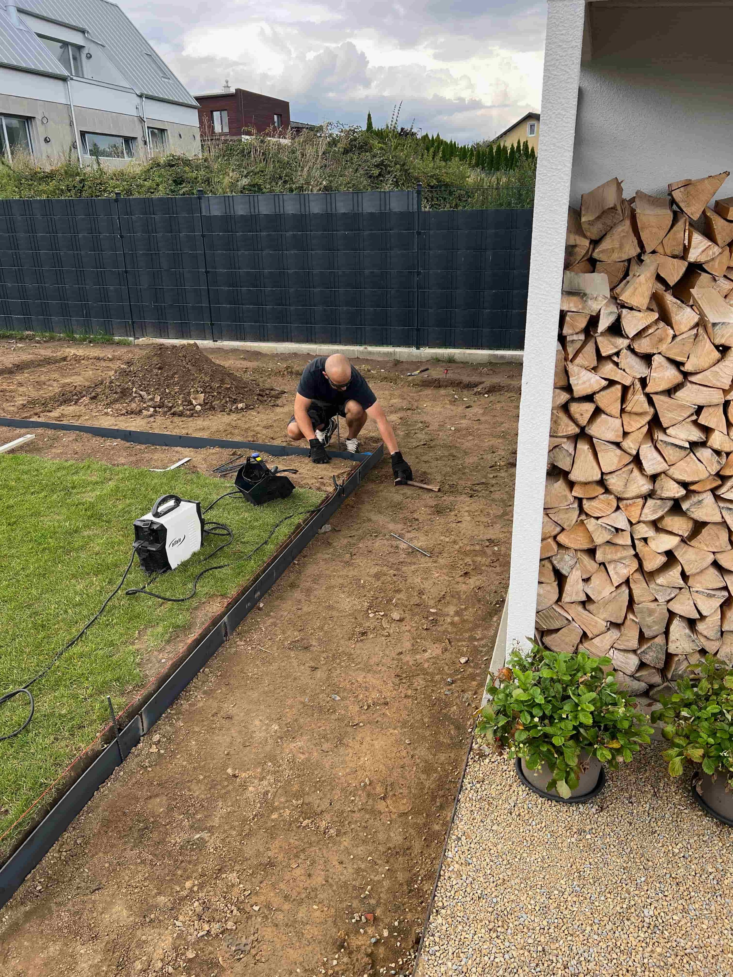 Práce na zahradě při realizaci – montáž kovového obrubníku u záhonu, v pozadí plot a dřevo na uskladnění.