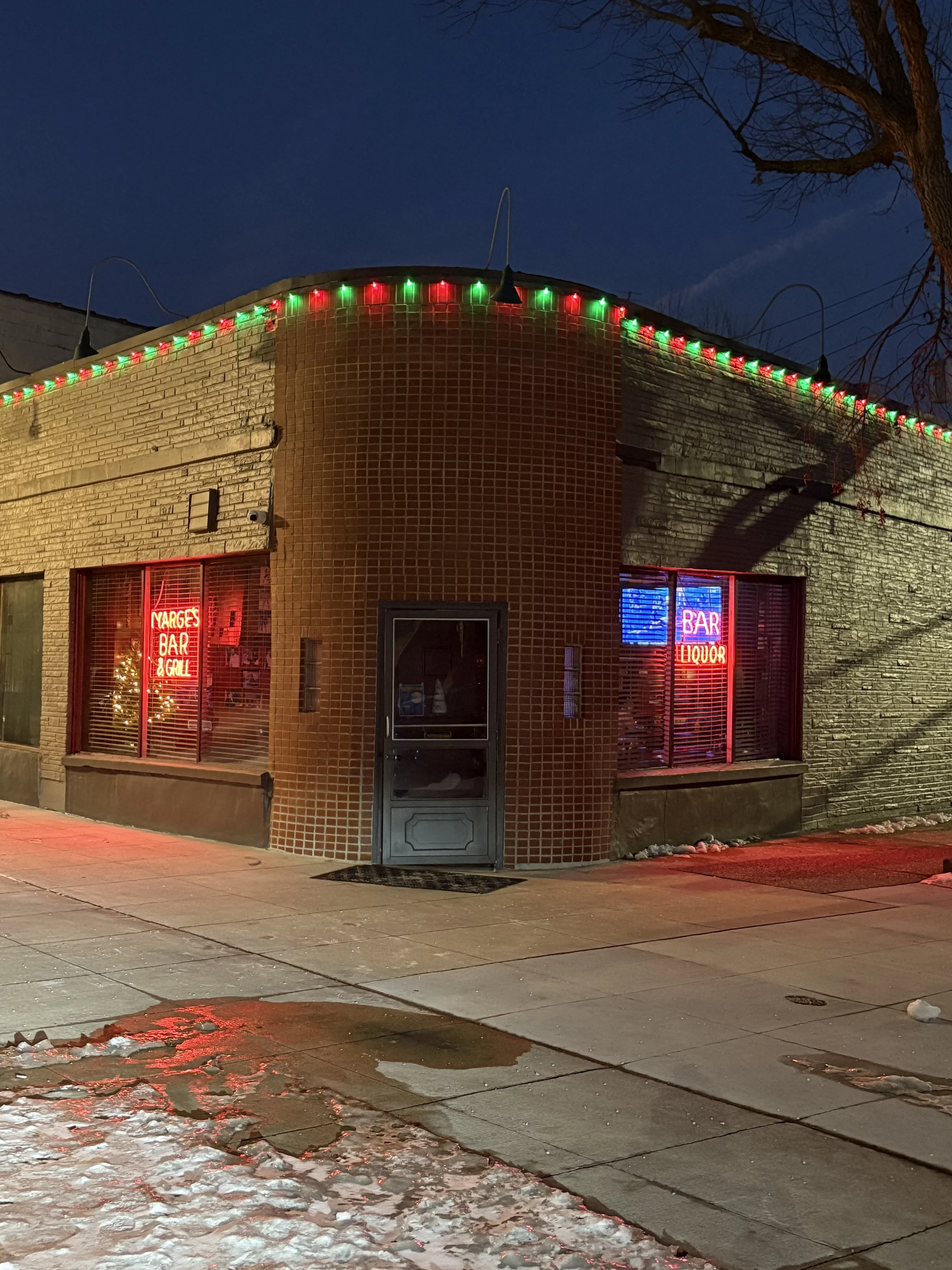Corner building with brick and stone walls, decorated with red and green Christmas lights along the roofline. Neon signs in the windows display 'MARGES BAR & GRILL' on the left and 'BAR LIQUOR' on the right. It is nighttime, with snow on the sidewalk and a clear sky.