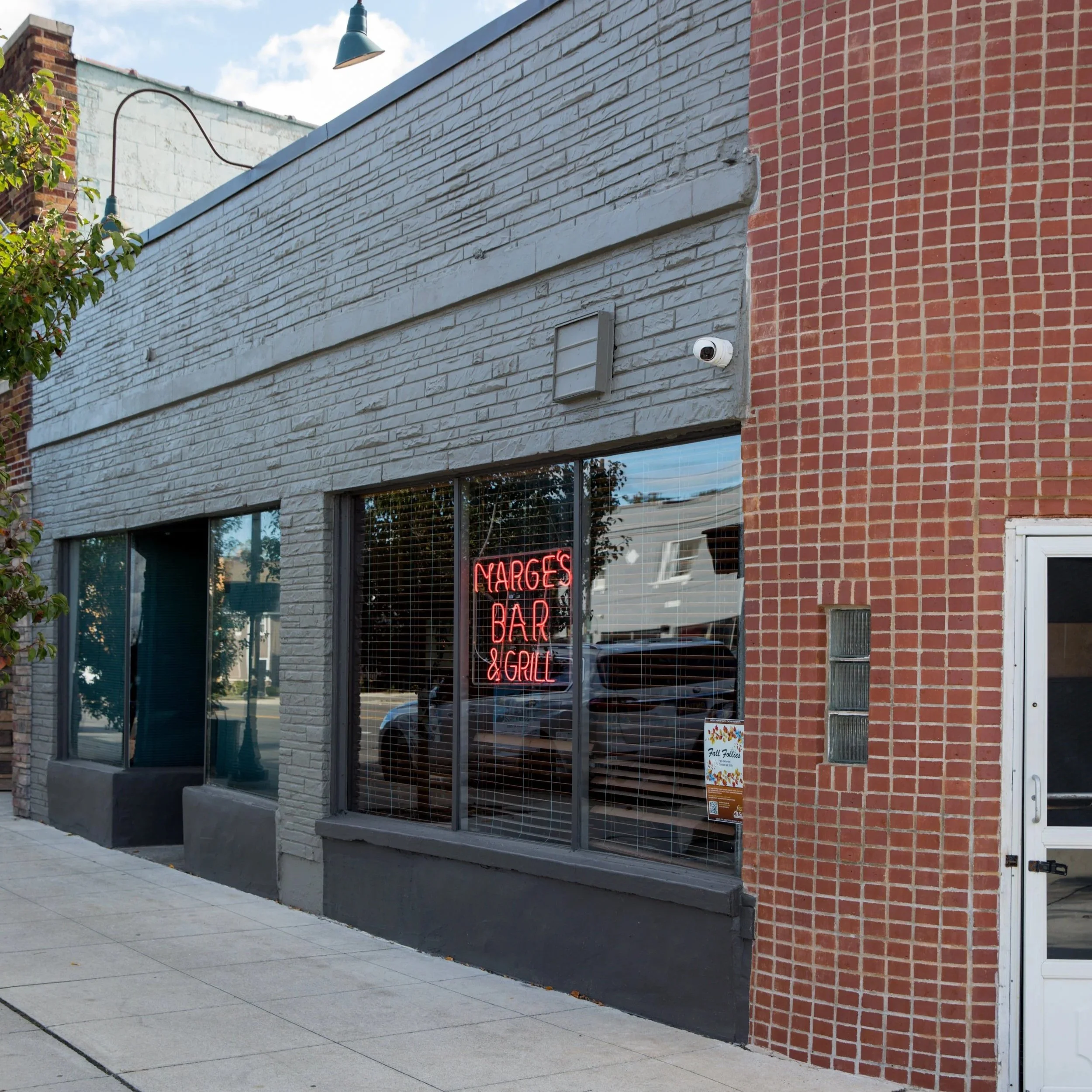 Exterior of a brick building with a large window displaying a neon sign that reads 'NARGES BAR & GRILL' and a security camera mounted on the wall.