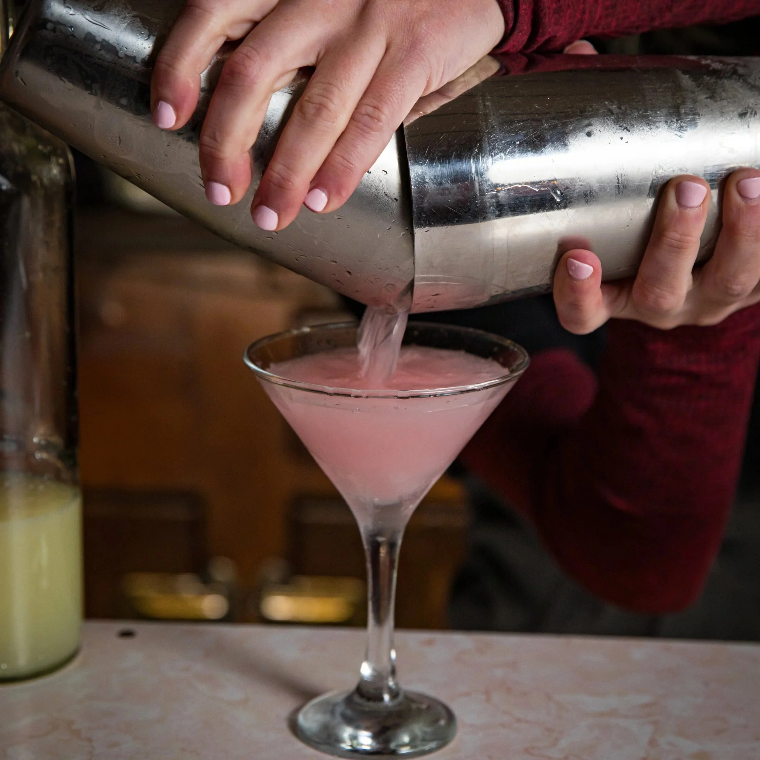 A person pours a pink cocktail from a cocktail shaker into a martini glass.