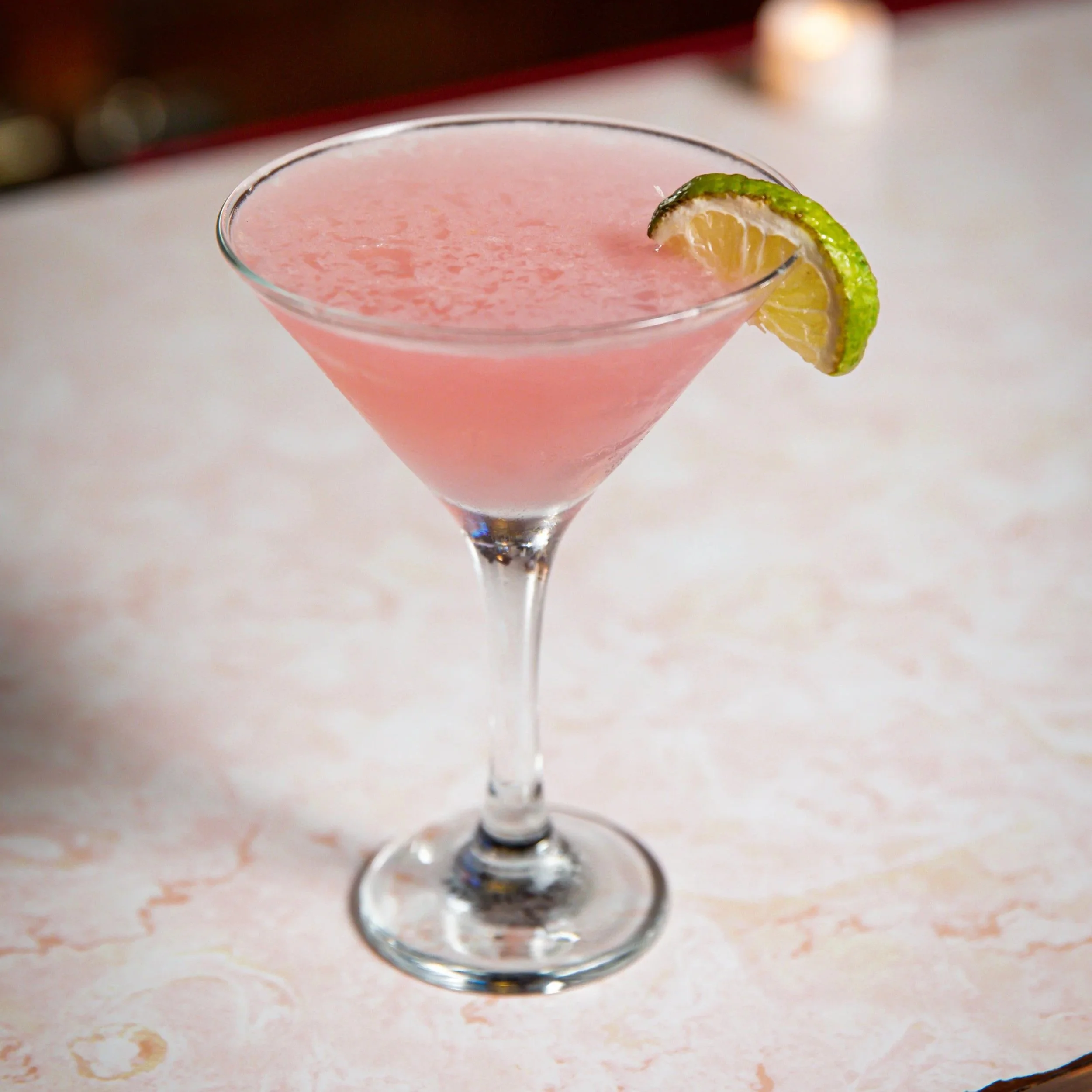 A pink cocktail in a martini glass with a lime wedge garnish.