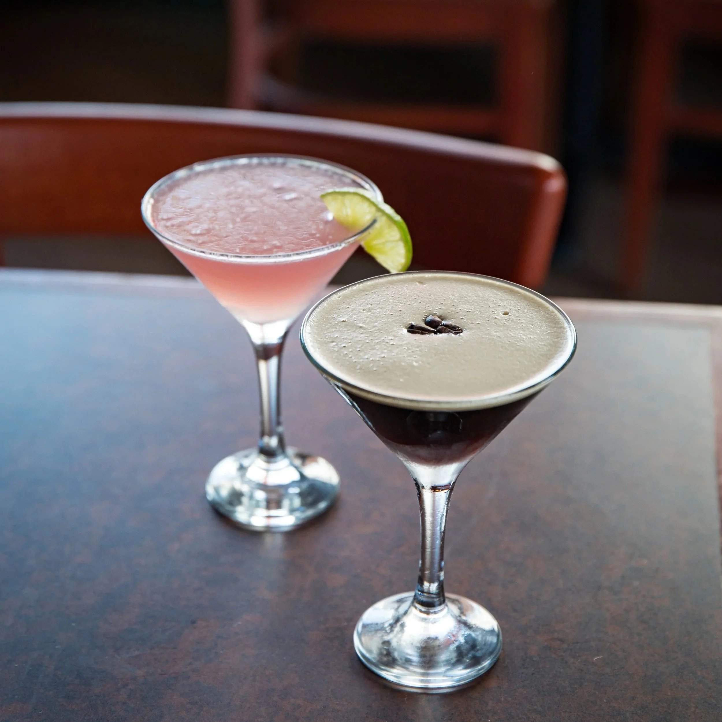 Two cocktails on a brown table, one is a pink drink garnished with lime and the other is a dark cocktail with cream or foam topping and coffee beans.