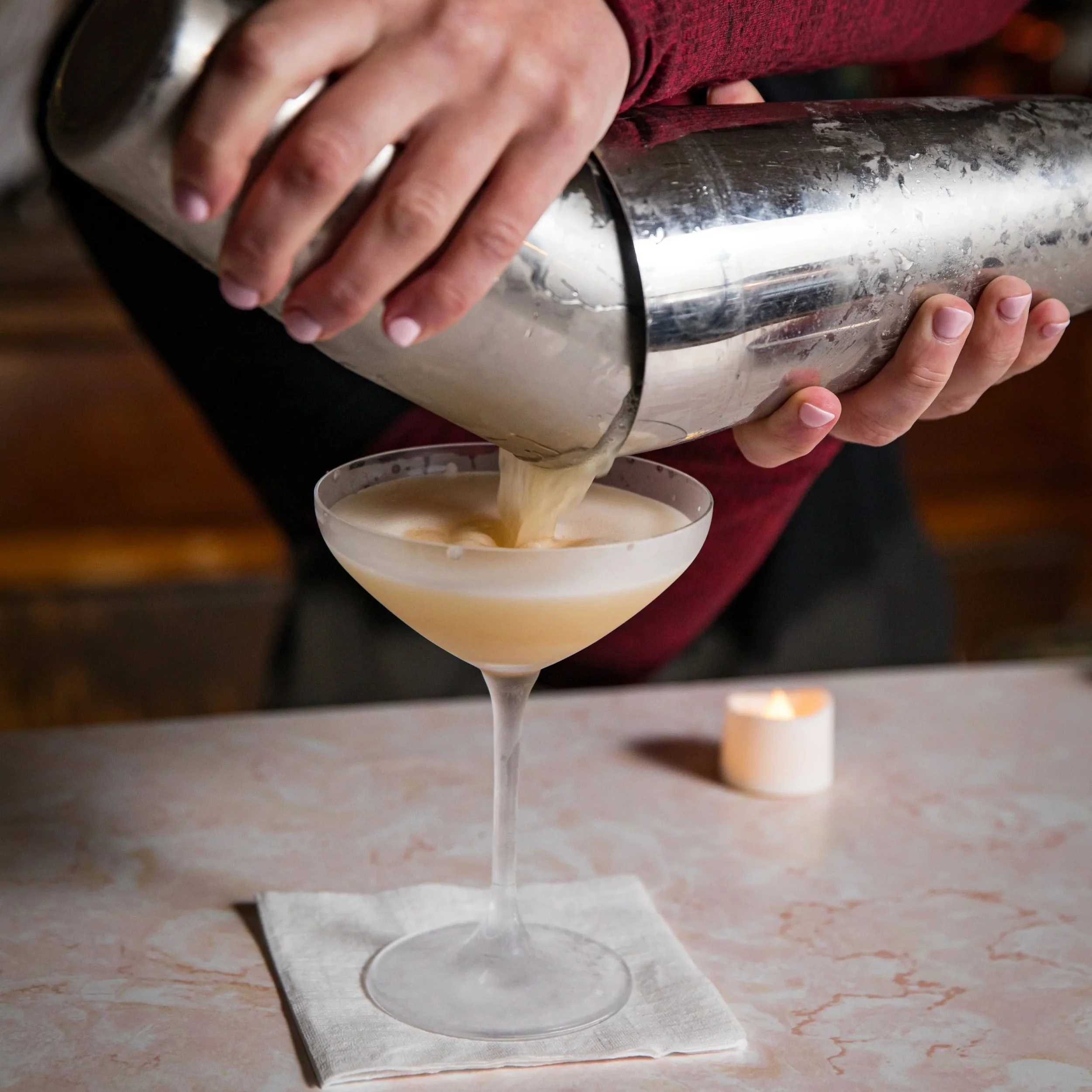 A bartender pours a creamy cocktail from a shaker into a coupe glass over a napkin, with a lit candle in the background.