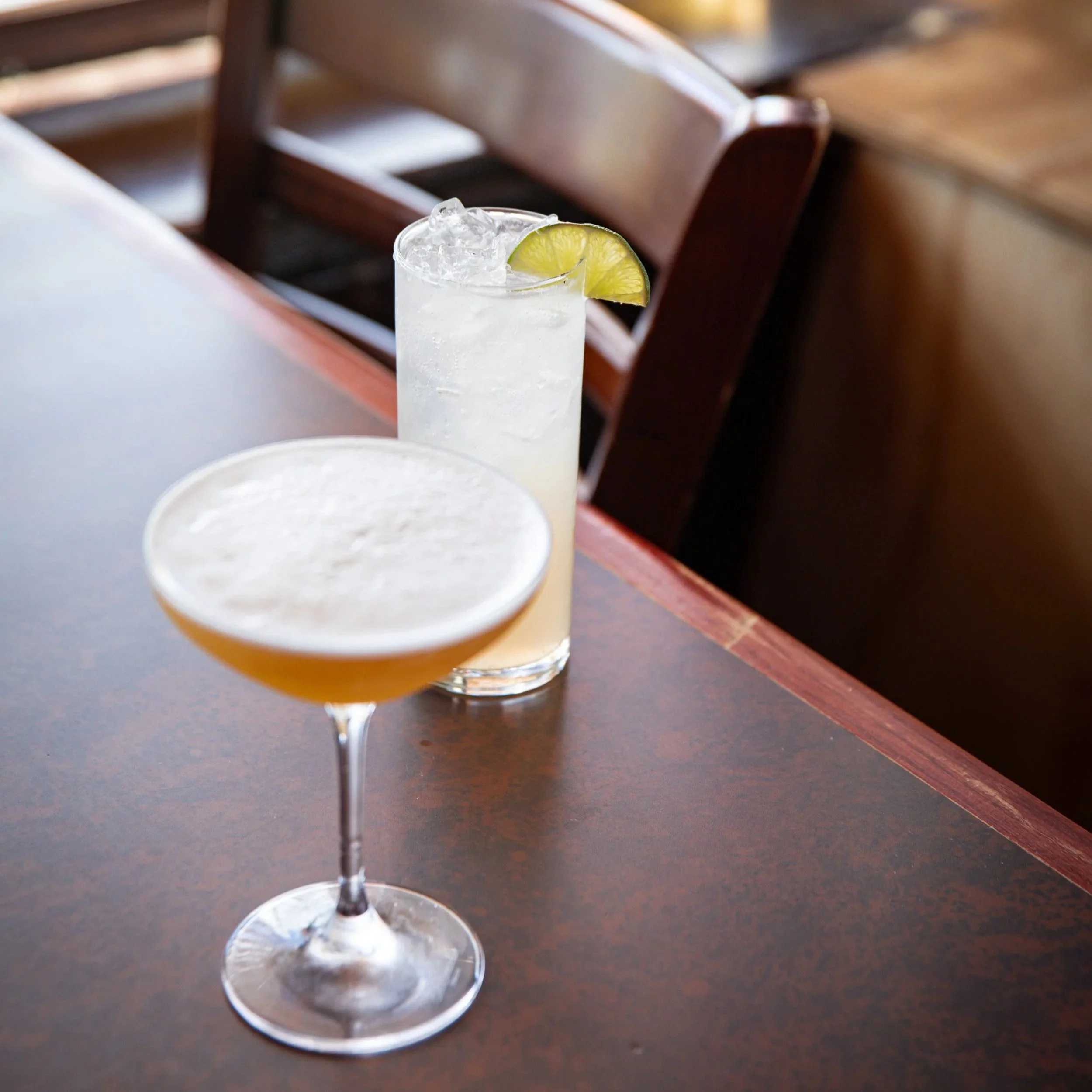 Two cocktails on a wooden bar counter, one in a coupe glass with a frothy top, and a tall glass with ice and a lime wedge.