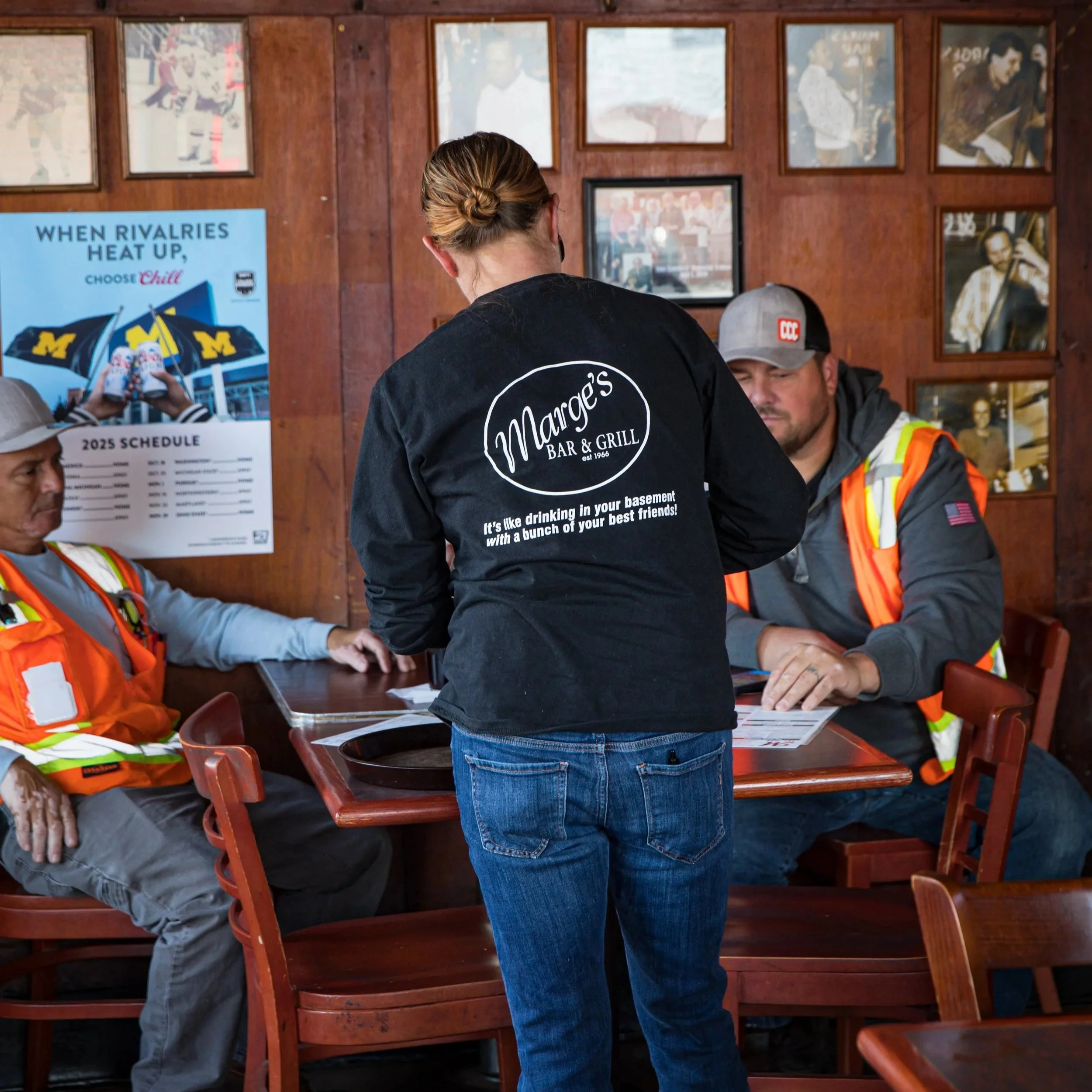 Inside a bar or restaurant with wooden walls, three people are sitting around a table. One woman, wearing a black t-shirt with white print, is standing and facing away from the camera. Two men, dressed in gray hoodies with orange safety vests, are si