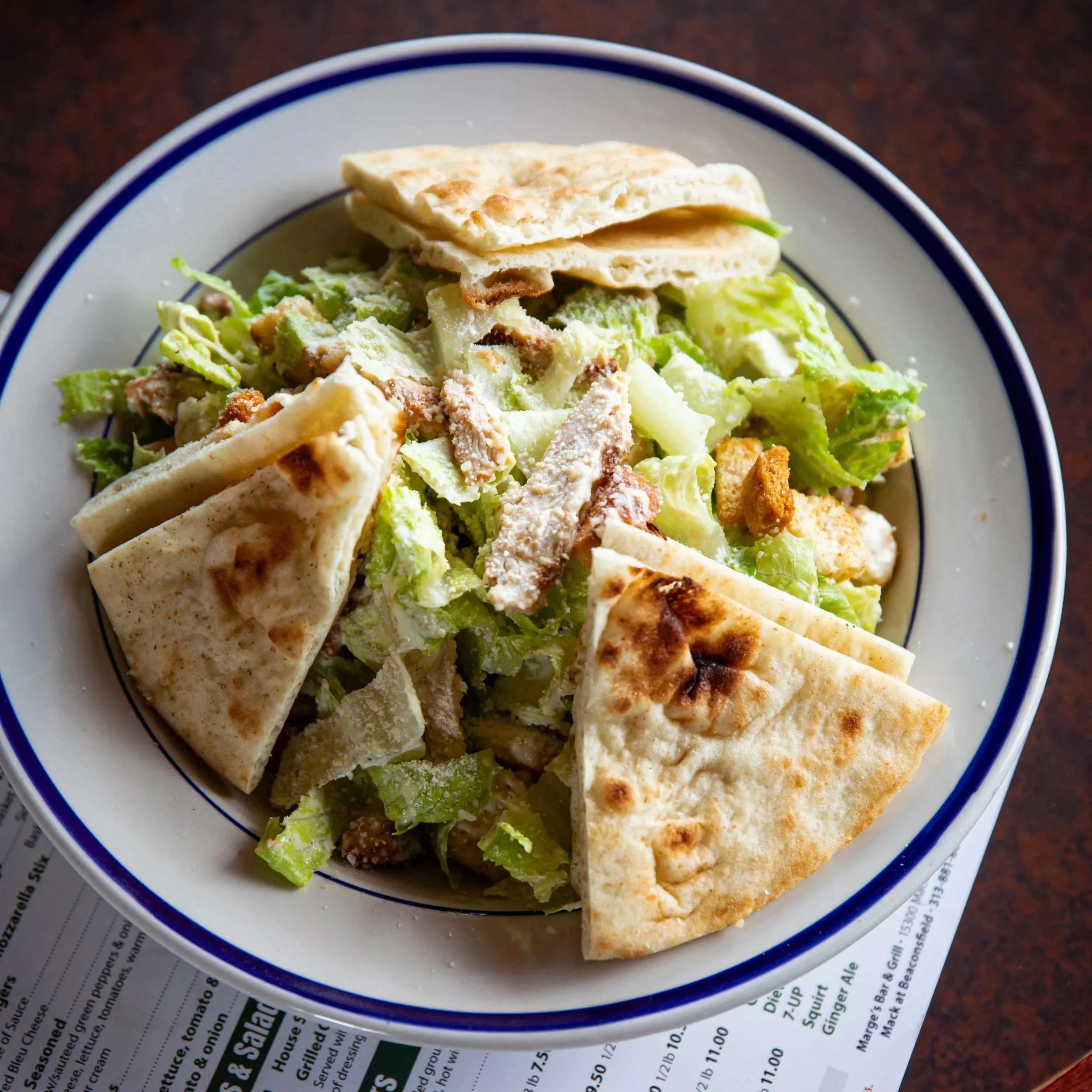 Caesar salad with grilled chicken, romaine lettuce, croutons, and grilled pita bread served in a white bowl with a blue rim.