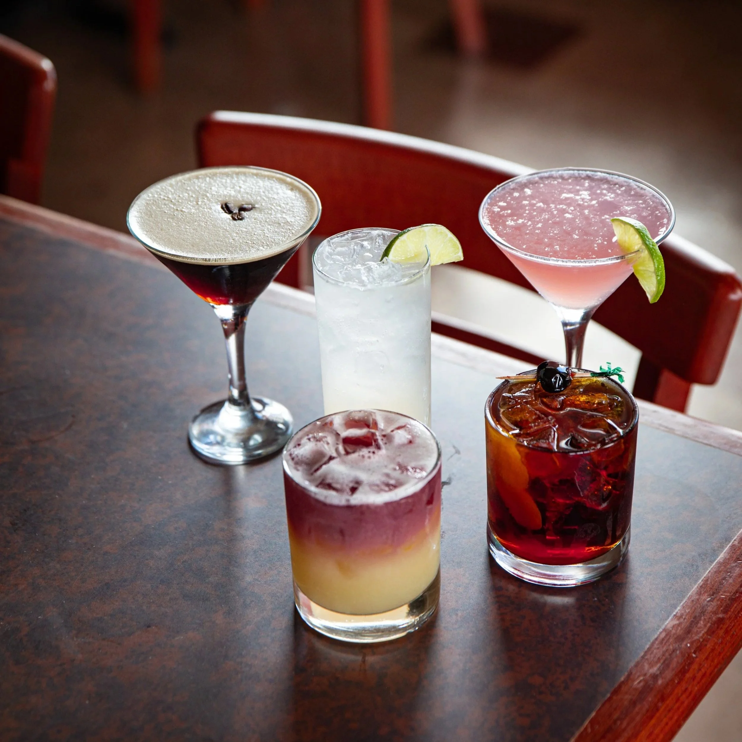 Five cocktails on a wooden table, including a martini with a dark red drink and olive, a margarita with a lime wedge, a pink cocktail with a lime wedge, a layered drink with yellow and purple layers, and a clear drink with lime. Red chairs are visibl