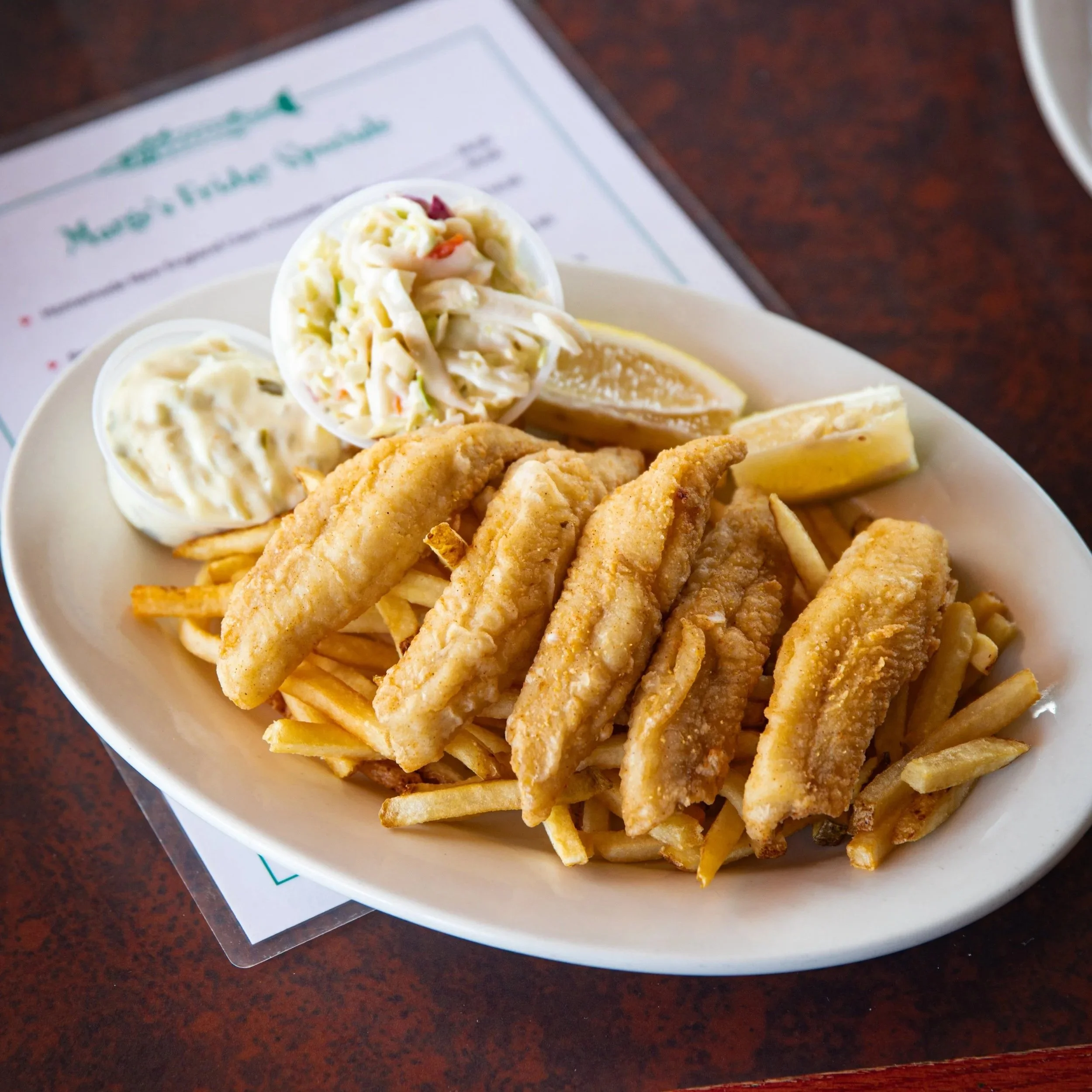 Plate of fried fish fillets, French fries, lemon wedges, and side dishes of coleslaw and tartar sauce.