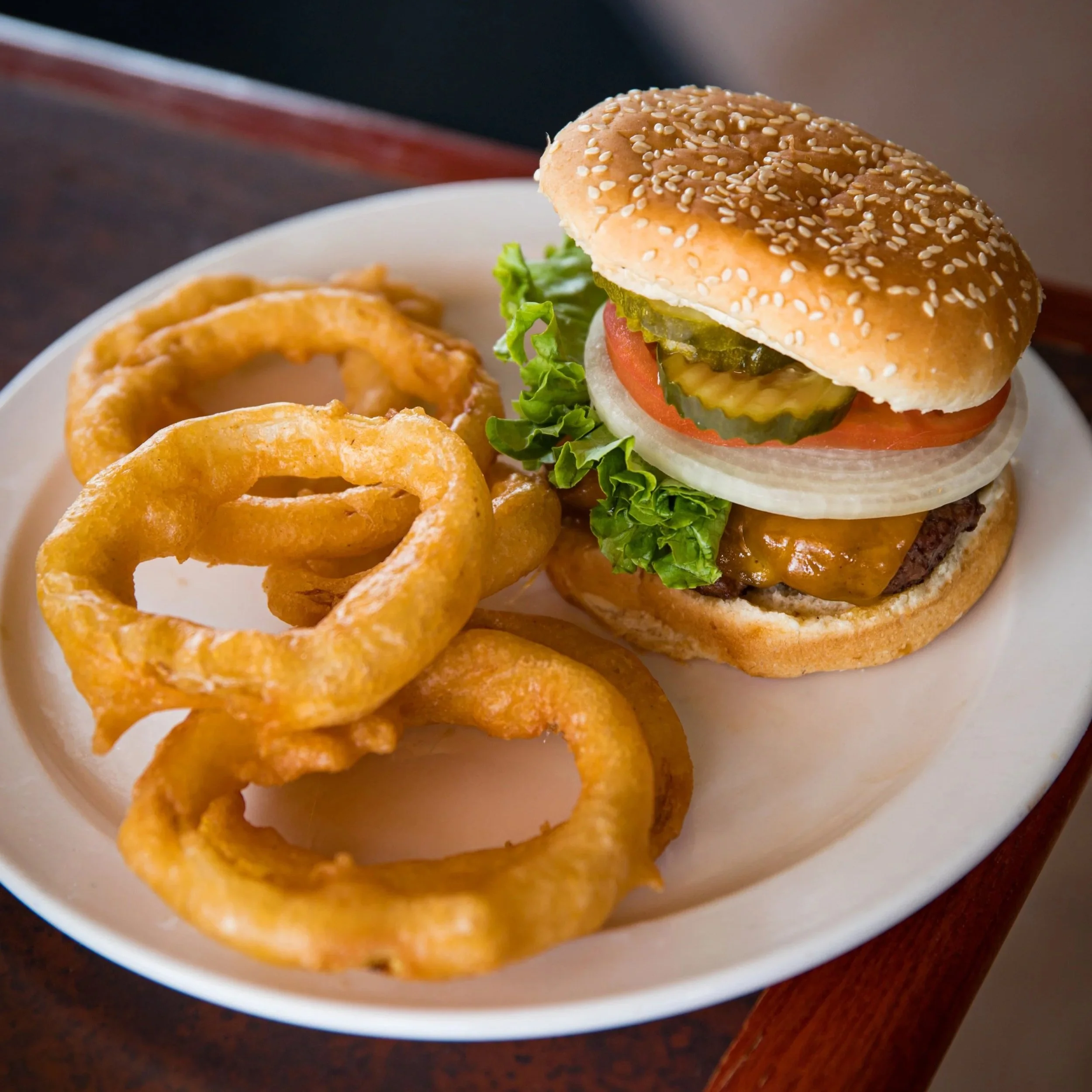 A cheeseburger with lettuce, tomato, pickles, onions, and a sesame seed bun, served with a side of curly onion rings on a white plate.