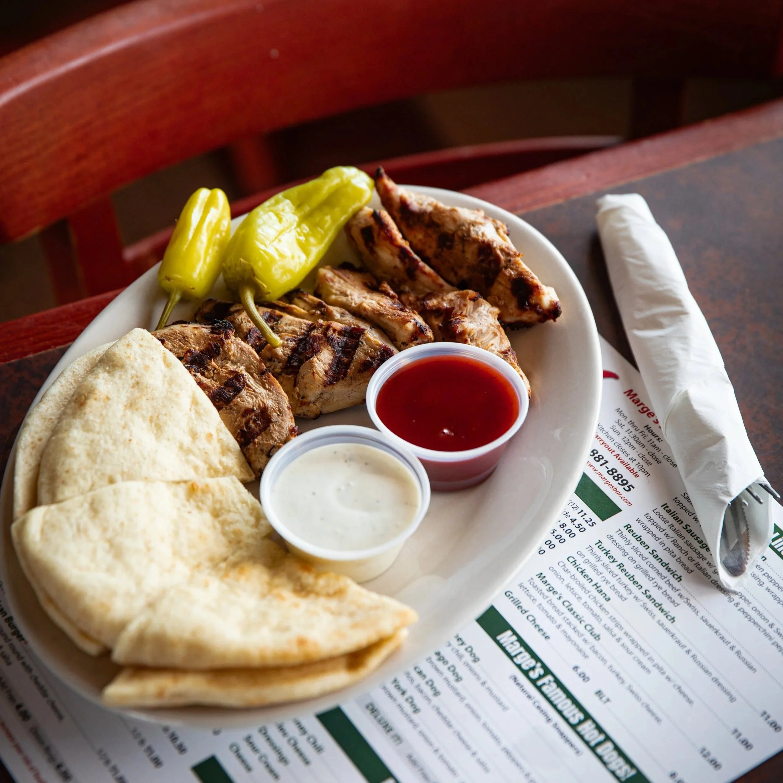 Plate with grilled chicken, pickled peppers, pita bread, and small containers of red sauce and white sauce, on top of a restaurant menu with a fork and napkin.