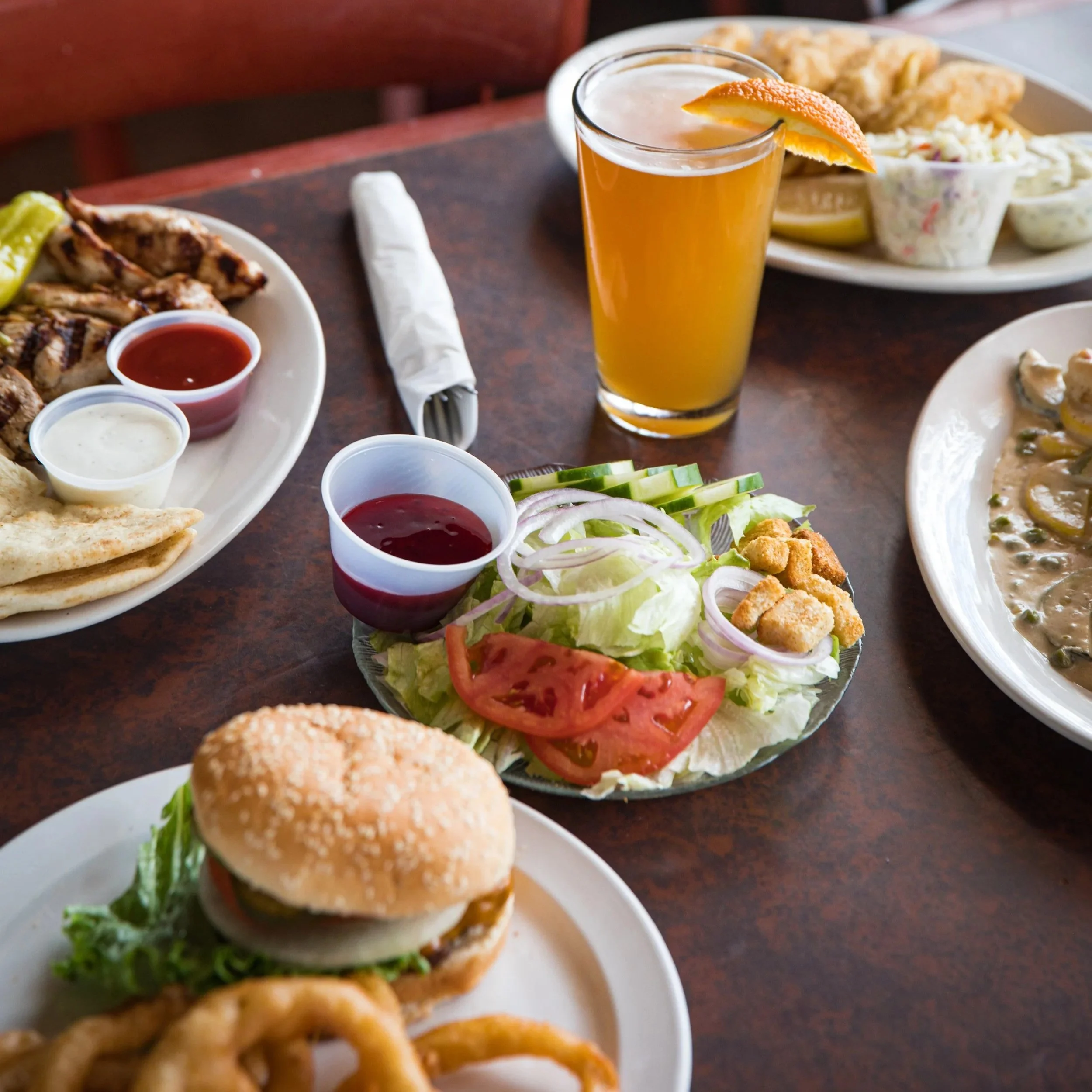 A table with various American lunch foods including a cheeseburger, a salad with tomatoes, onions, and cucumbers, chicken wings, fried fish, and a glass of orange juice with an orange slice garnishing the rim.