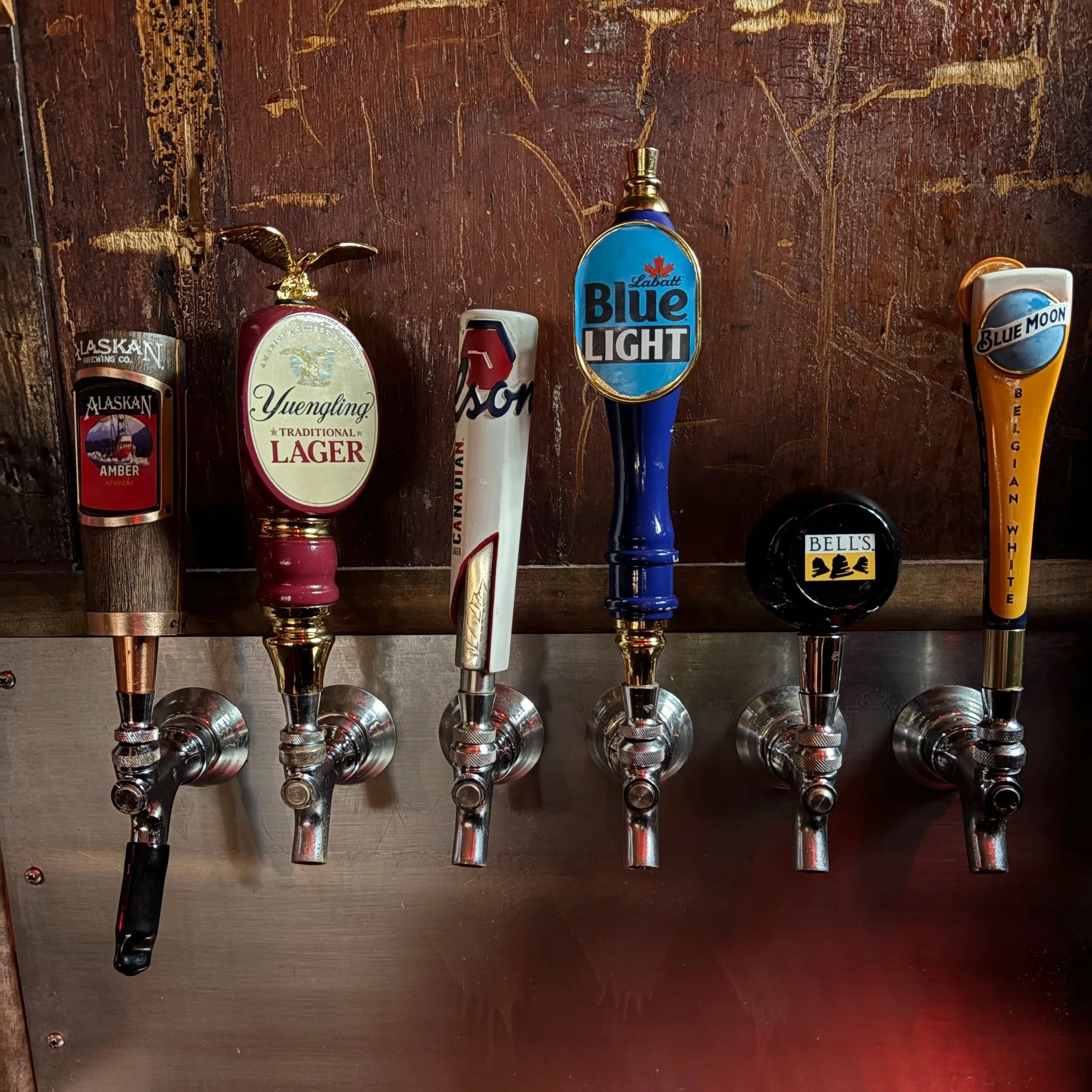 Six beer tap handles on a wall, labeled from left to right: Alaskan Amber, Yuengling Traditional Lager, Canadian, Labatt Blue Light, Bell's, Blue Moon Belgian White.