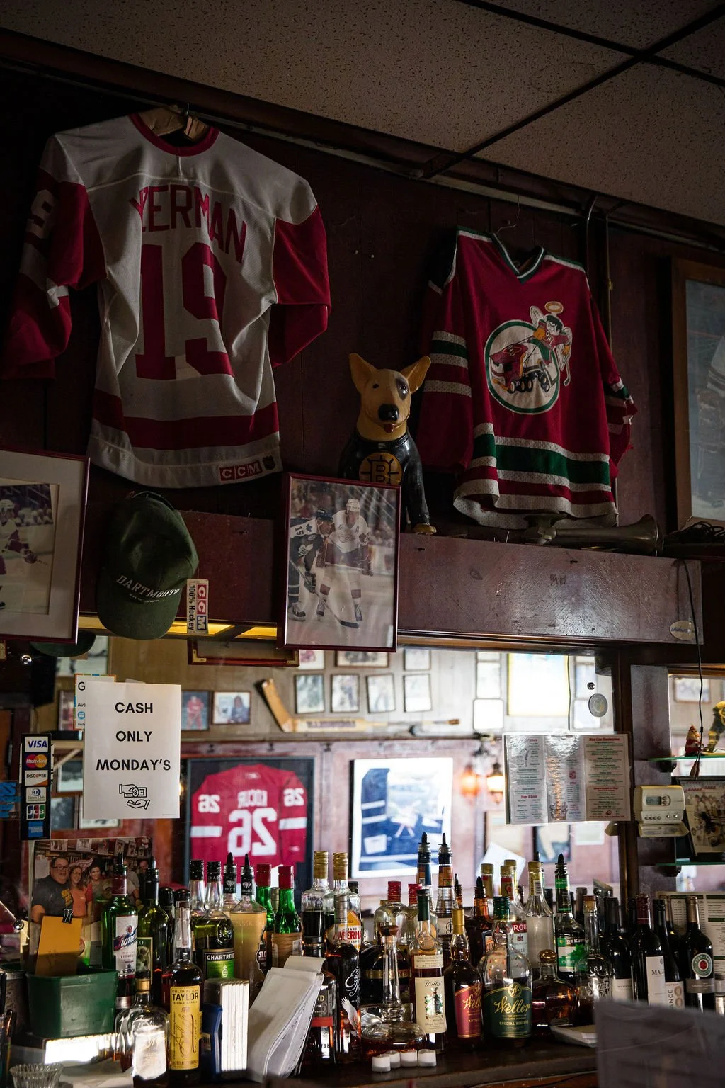 Hockey memorabilia displayed on a wooden wall, including jerseys and photos, with a bar counter filled with various bottles of alcohol in the foreground.