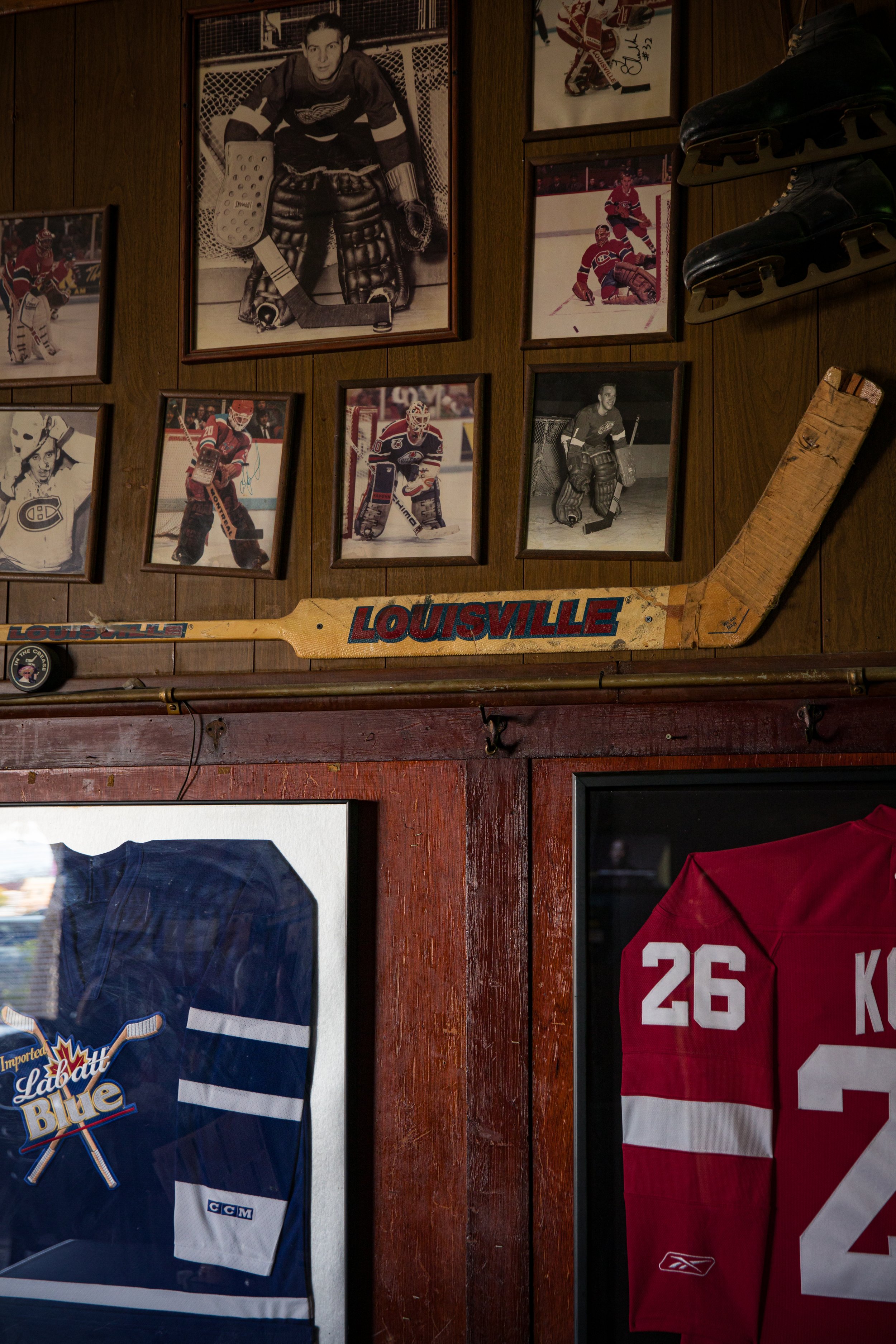Collection of framed hockey photos, old wooden hockey stick with 'LOUISVILLE' written on it, hockey shoes on the wall, part of a hockey jersey, and a display of hockey memorabilia.