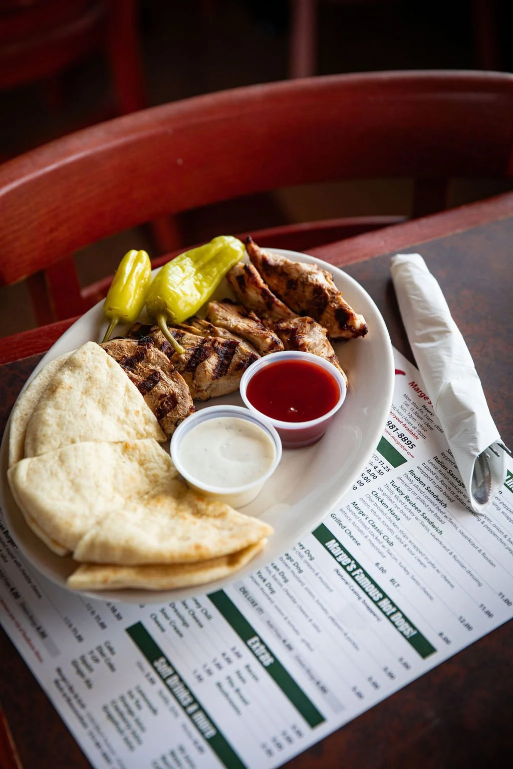 Plate of grilled chicken, green peppers, flatbread, and two dipping sauces on a restaurant table with a menu and napkin.