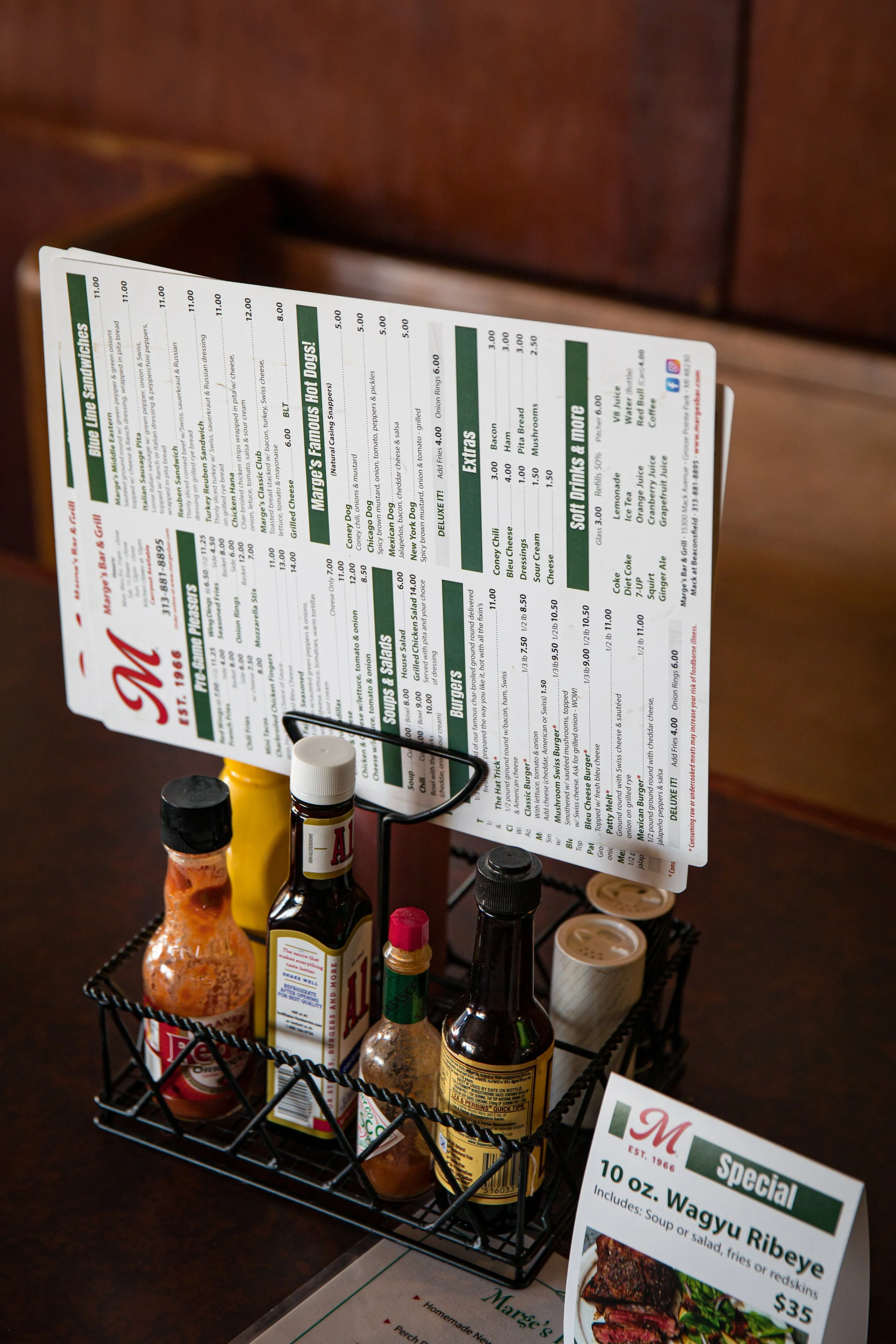 A tabletop restaurant setup with a metal caddy holding condiments and two salt shakers. A menu is propped up on a stand behind it, and a small special menu card advertises a 10 oz. Wagyu Ribeye steak for $35.