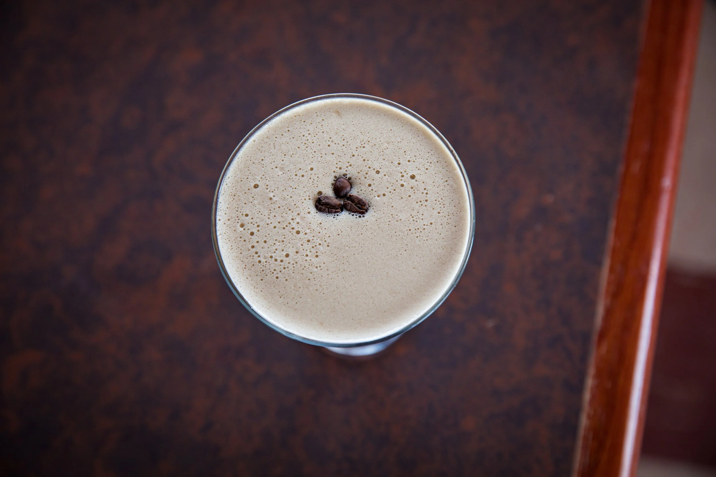 A top-down view of a glass of coffee with a few coffee beans on top, placed on a dark wooden surface.