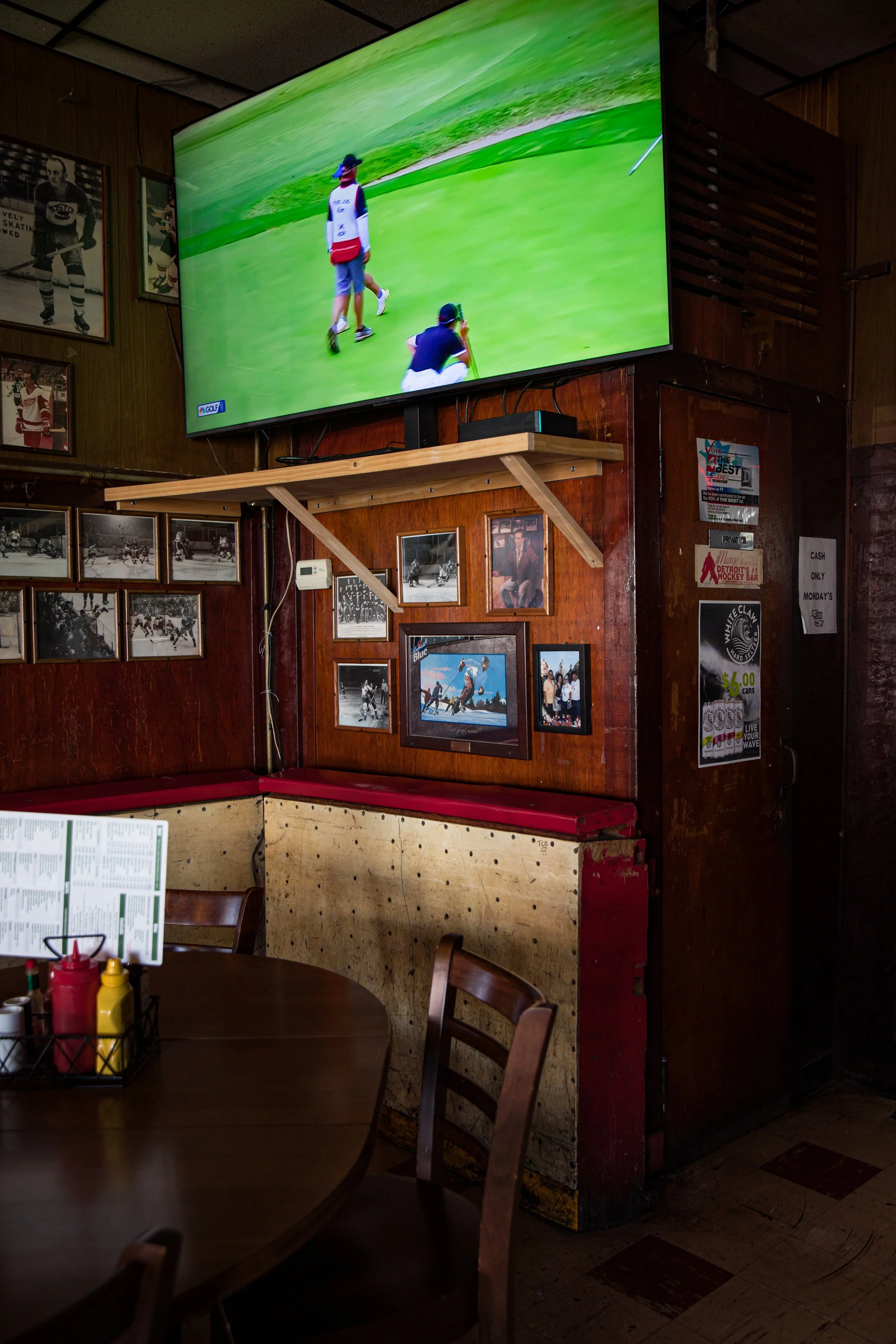 Photo of a restaurant or bar interior with a large television screen showing a golf game. The wall is decorated with black and white and color photographs, and there are wooden chairs and tables with condiments.