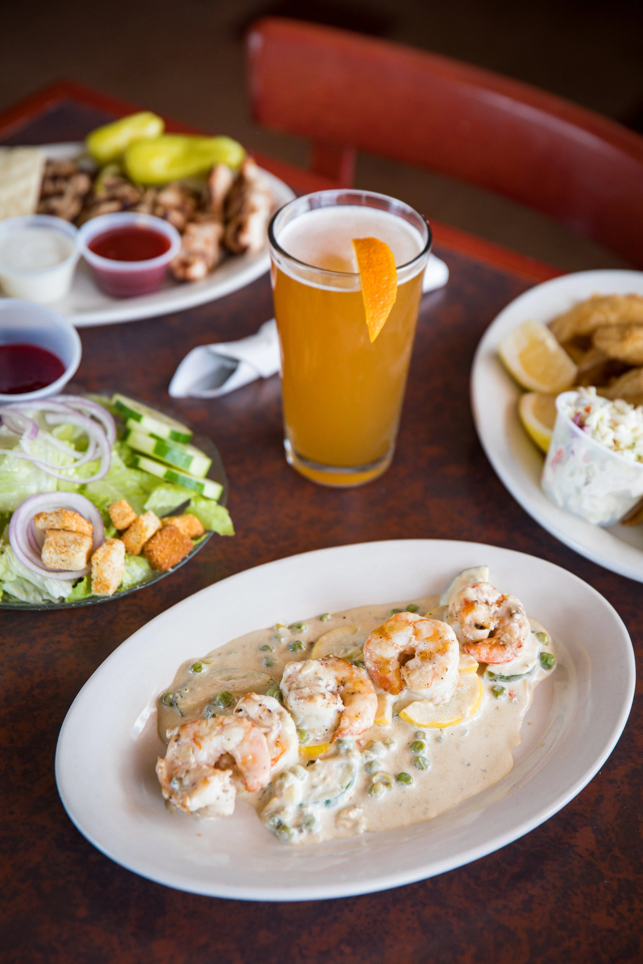 Shrimp with creamy white sauce, green peas, and lemon slices on a white plate, a salad with lettuce, cucumber, onions, and croutons, a glass of orange juice garnished with an orange peel, and other plates of food including fried chicken and dipping s