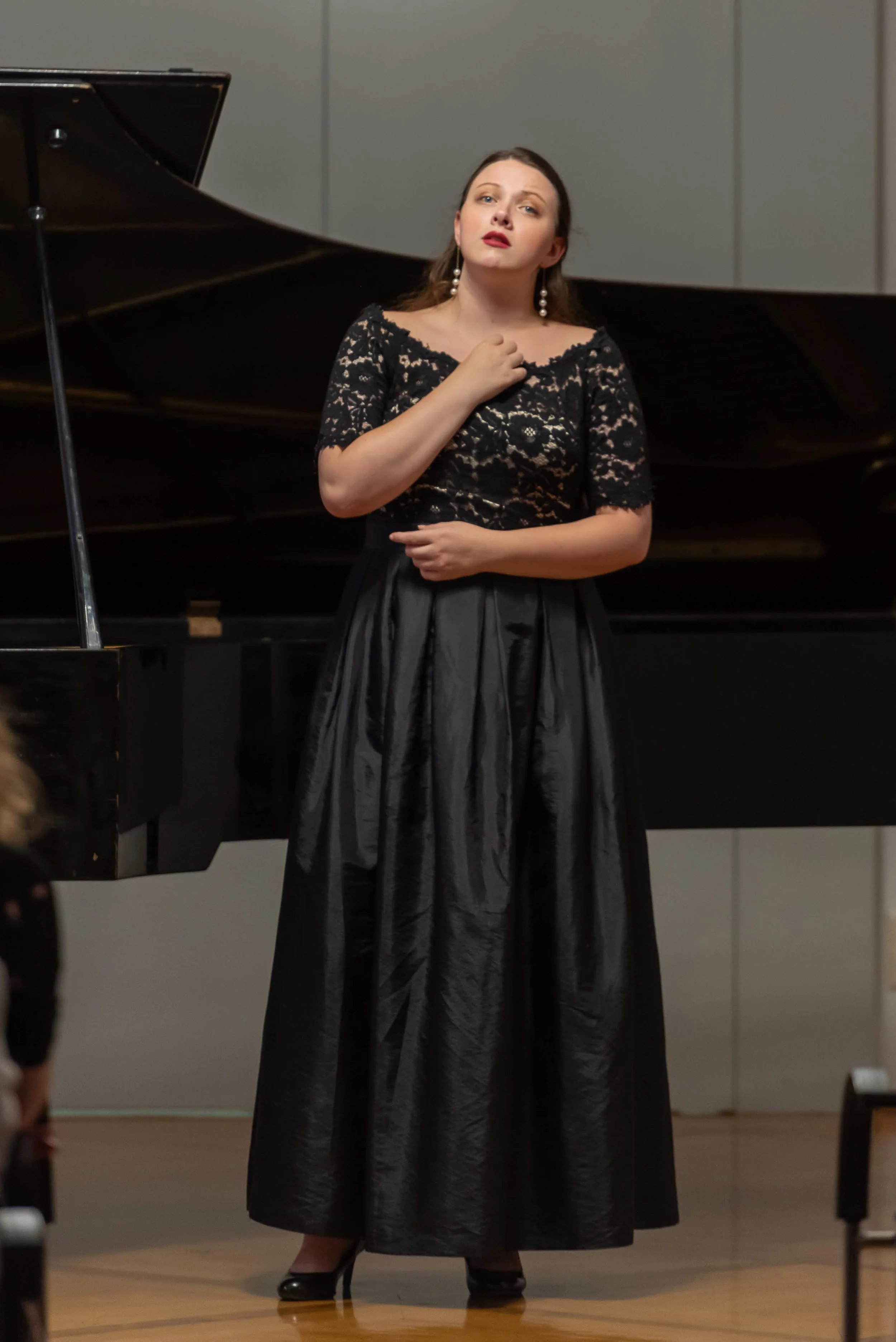 A woman, soprano Sarah Nalty, in a black lace top and a long black satin skirt stands in front of a grand piano on a stage, performing with a serious expression.
