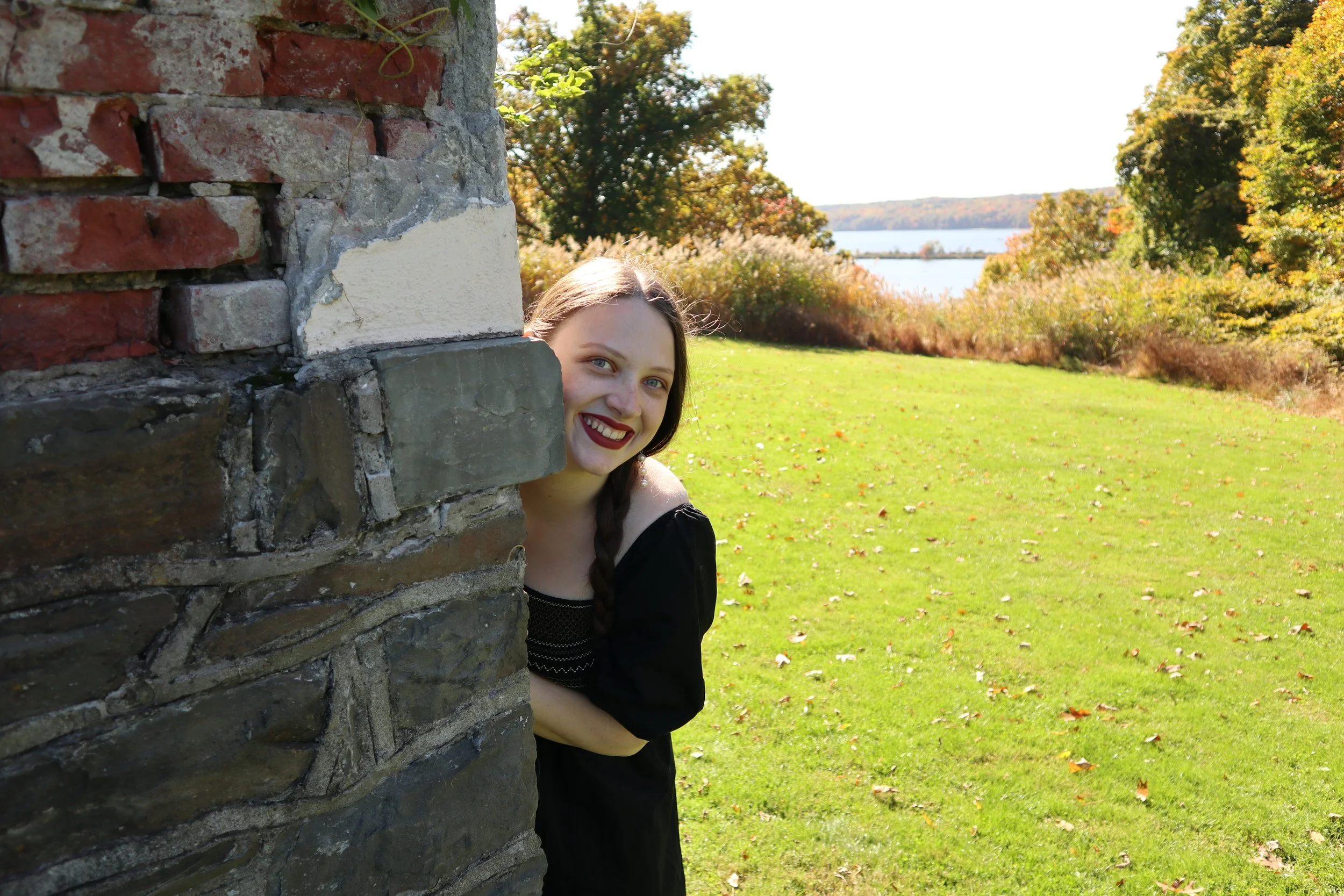 A woman, soprano Sarah Nalty, with long brown hair and red lipstick peeks out from behind a stone brick wall, smiling. She is outdoors on a sunny day with green grass, trees with fall foliage, and a body of water in the background.