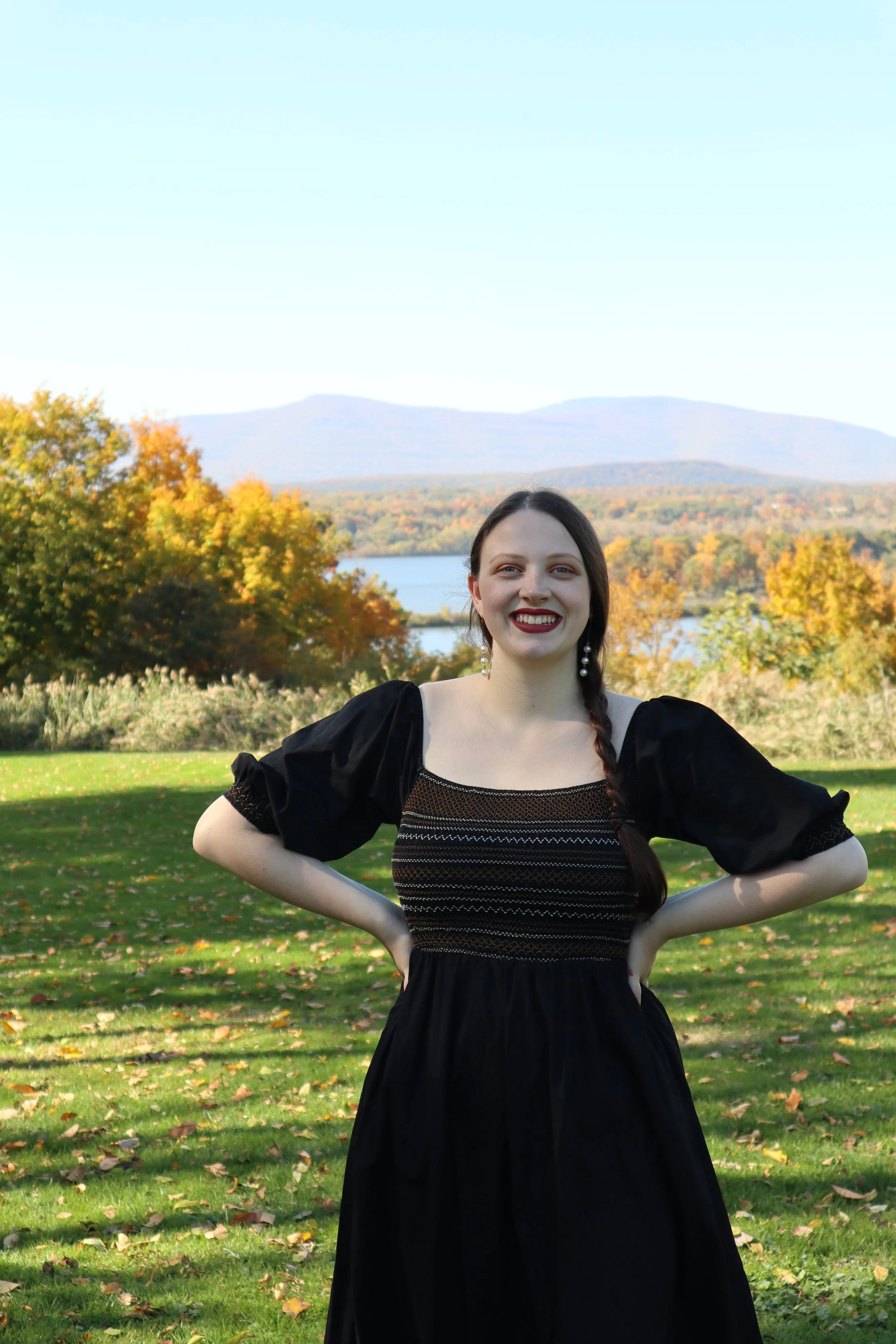 A woman, soprano Sarah Nalty, with dark hair in a braid, wearing a black dress with puffed sleeves, smiling, standing outdoors on grass with autumn trees, a lake, and mountains in the background.