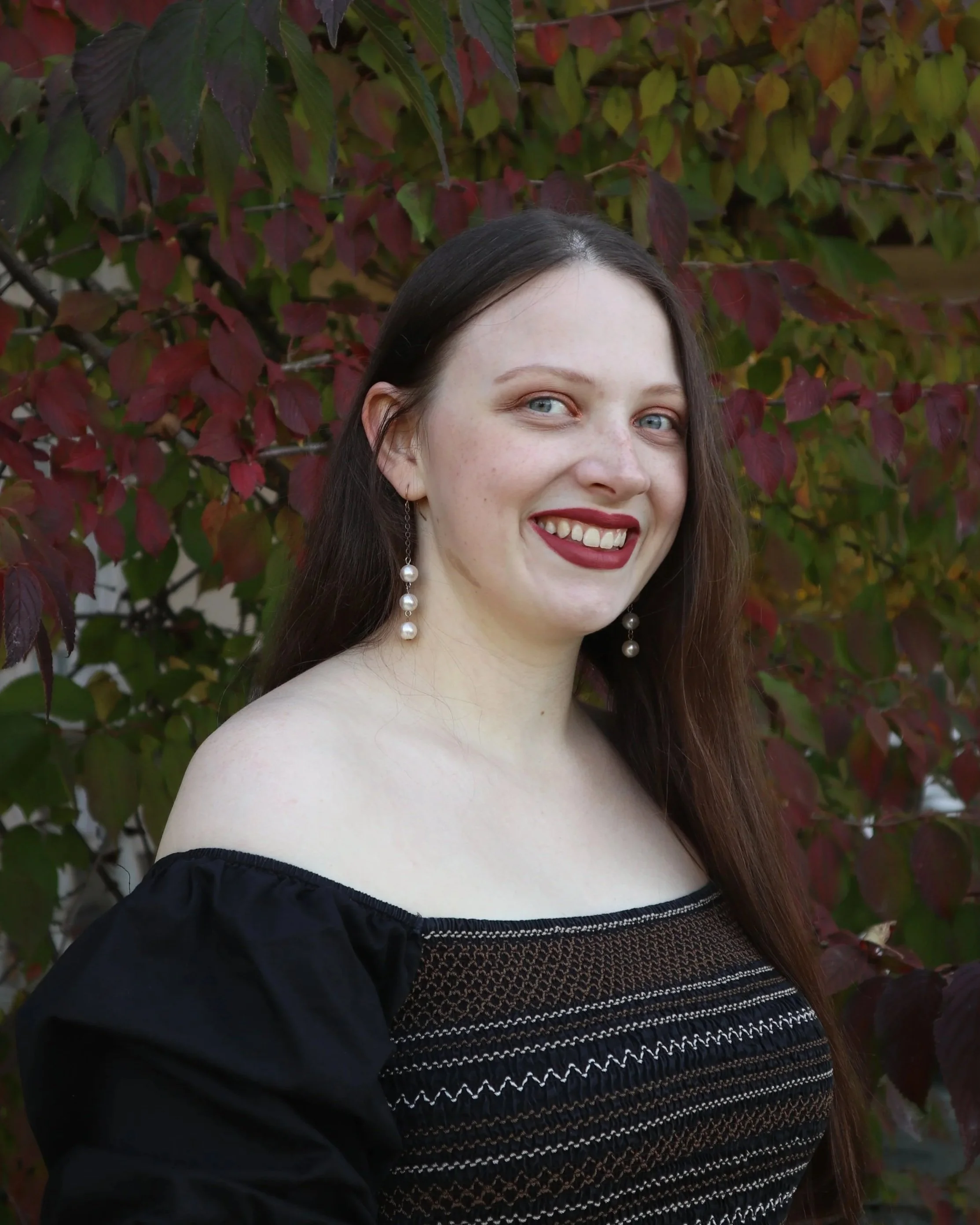 A young woman, soprano Sarah Nalty, with long brown hair and blue eyes smiling, wearing a black off-shoulder top with striped details and pearl earrings, standing in front of a background of red and green leaves.
