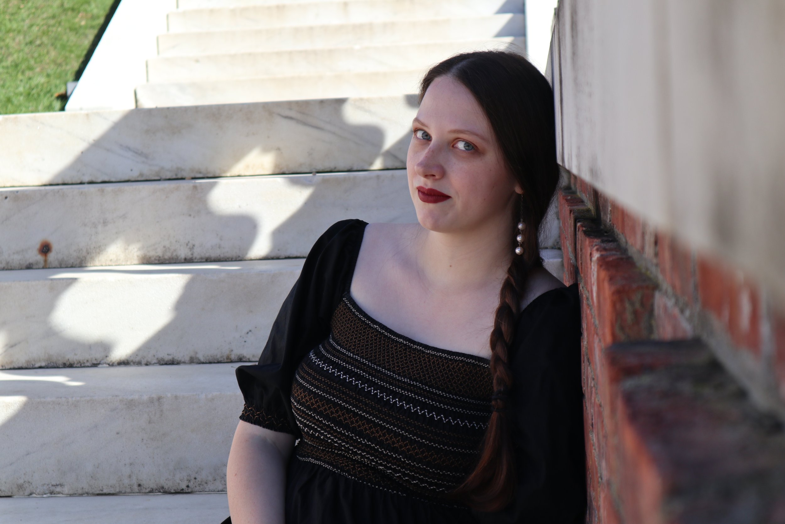 A woman, soprano Sarah Nalty, with dark hair, wearing a black dress with intricate embroidery, black jacket, and pearl earrings, leaning against a brick wall with white stairs and green grass in the background.