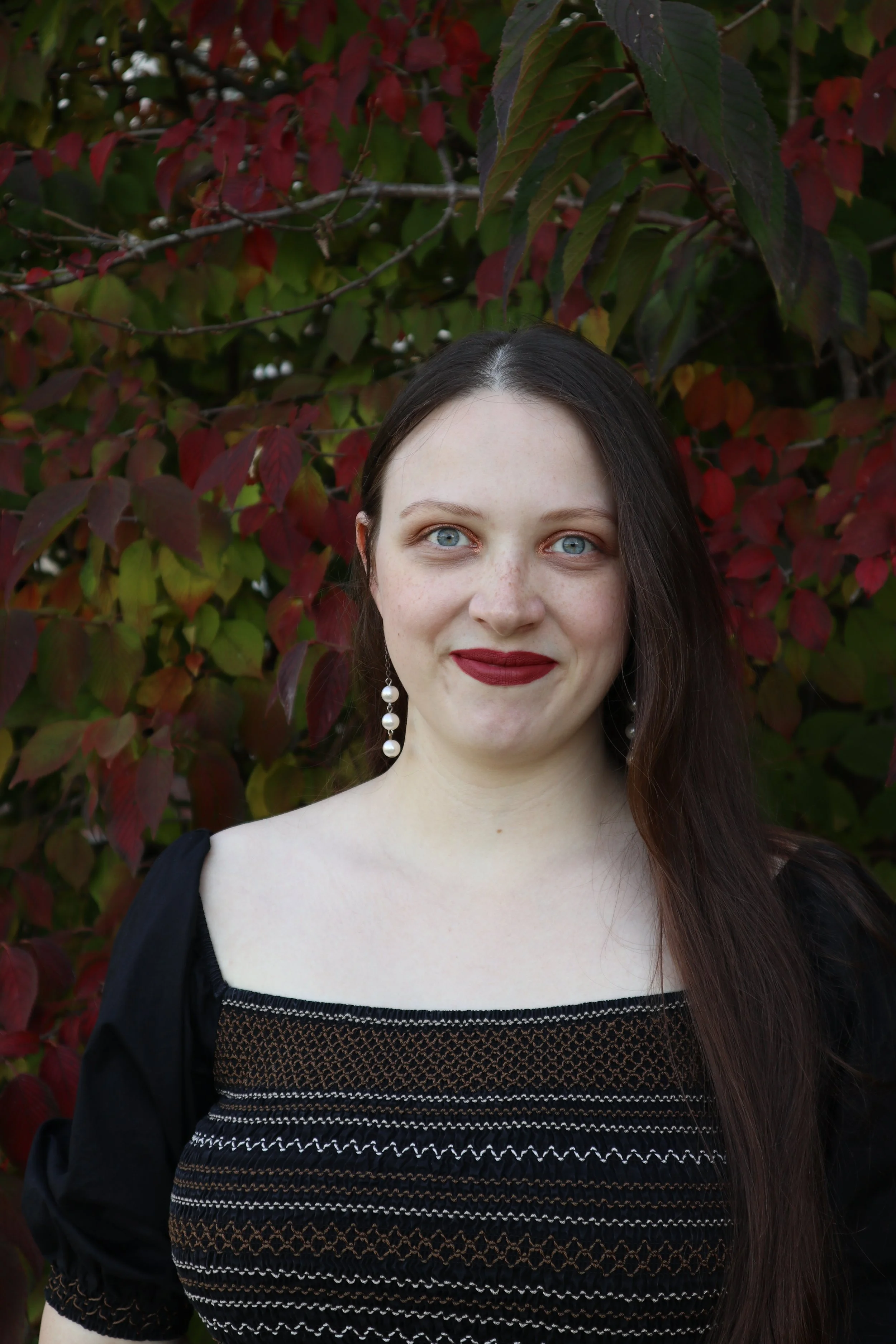 Portrait of a woman, soprano Sarah Nalty, with long brown hair, blue eyes, dressed in a black top with patterned embroidery, standing in front of a red and green foliage background.