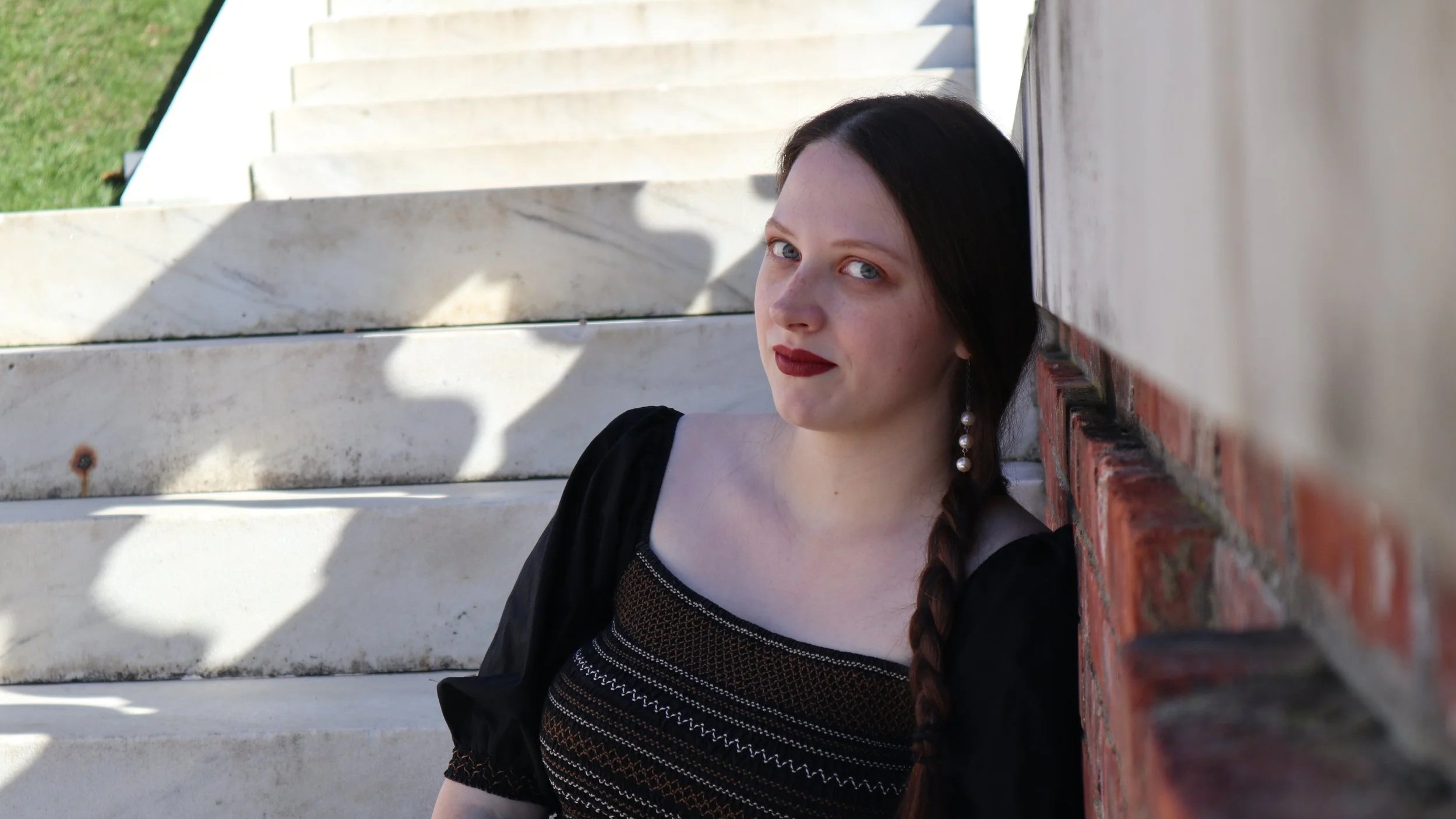 A woman, soprano Sarah Nalty, with dark hair in a braid, wearing red lipstick and pearl earrings, sitting on white marble stairs and leaning against a brick wall, looking at the camera.