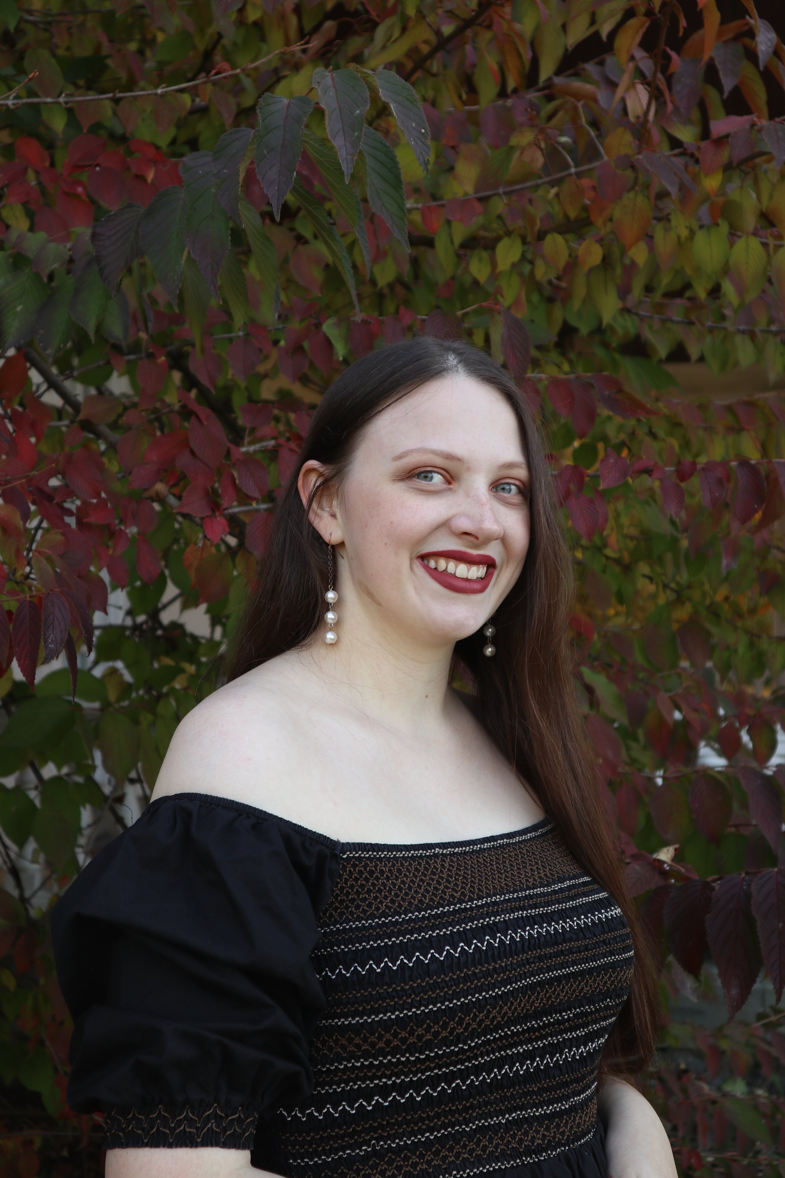 A young woman, soprano Sarah Nalty, with long dark hair, wearing an off-the-shoulder black dress with embroidery, pearl earrings, and dark lipstick, standing outdoors in front of a background of red and green leaves.