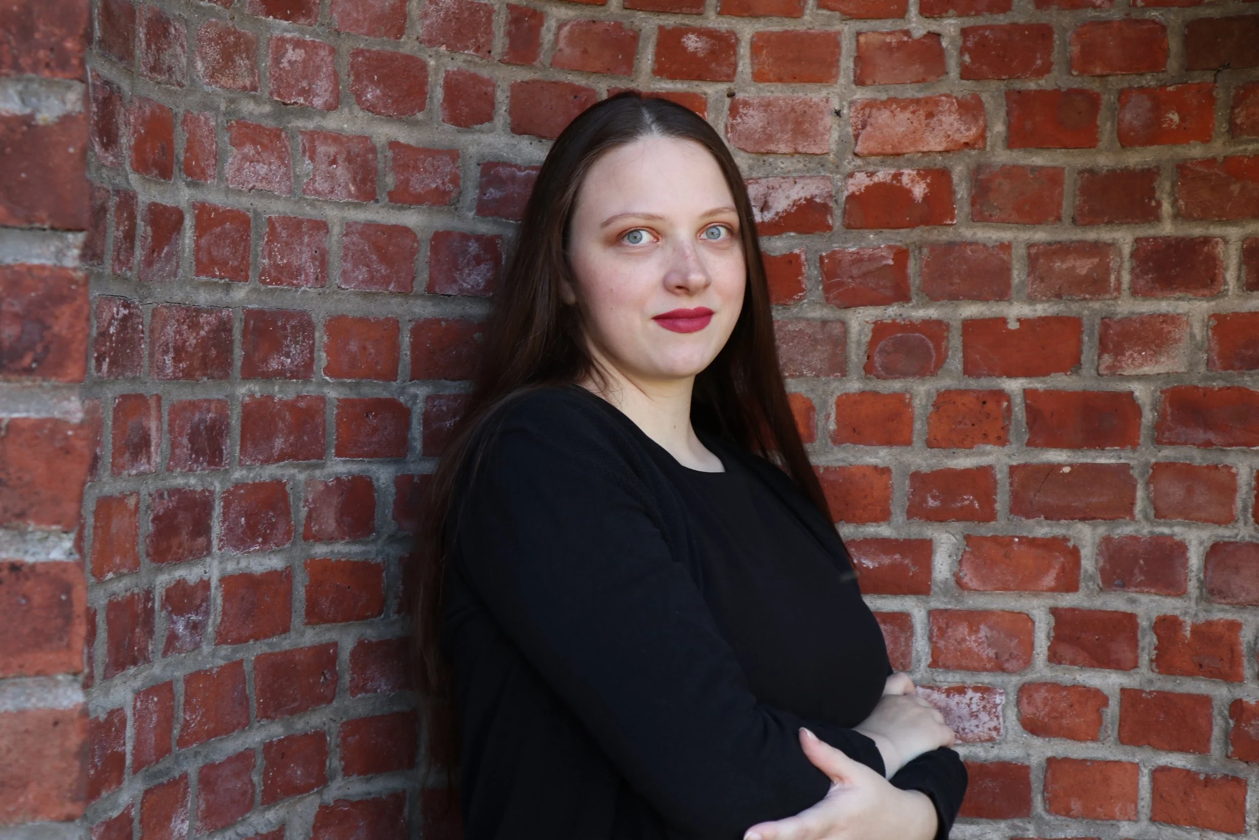 A woman, soprano Sarah Nalty, with long dark hair, wearing a black top, standing against a red brick wall, with her arms crossed and looking at the camera.