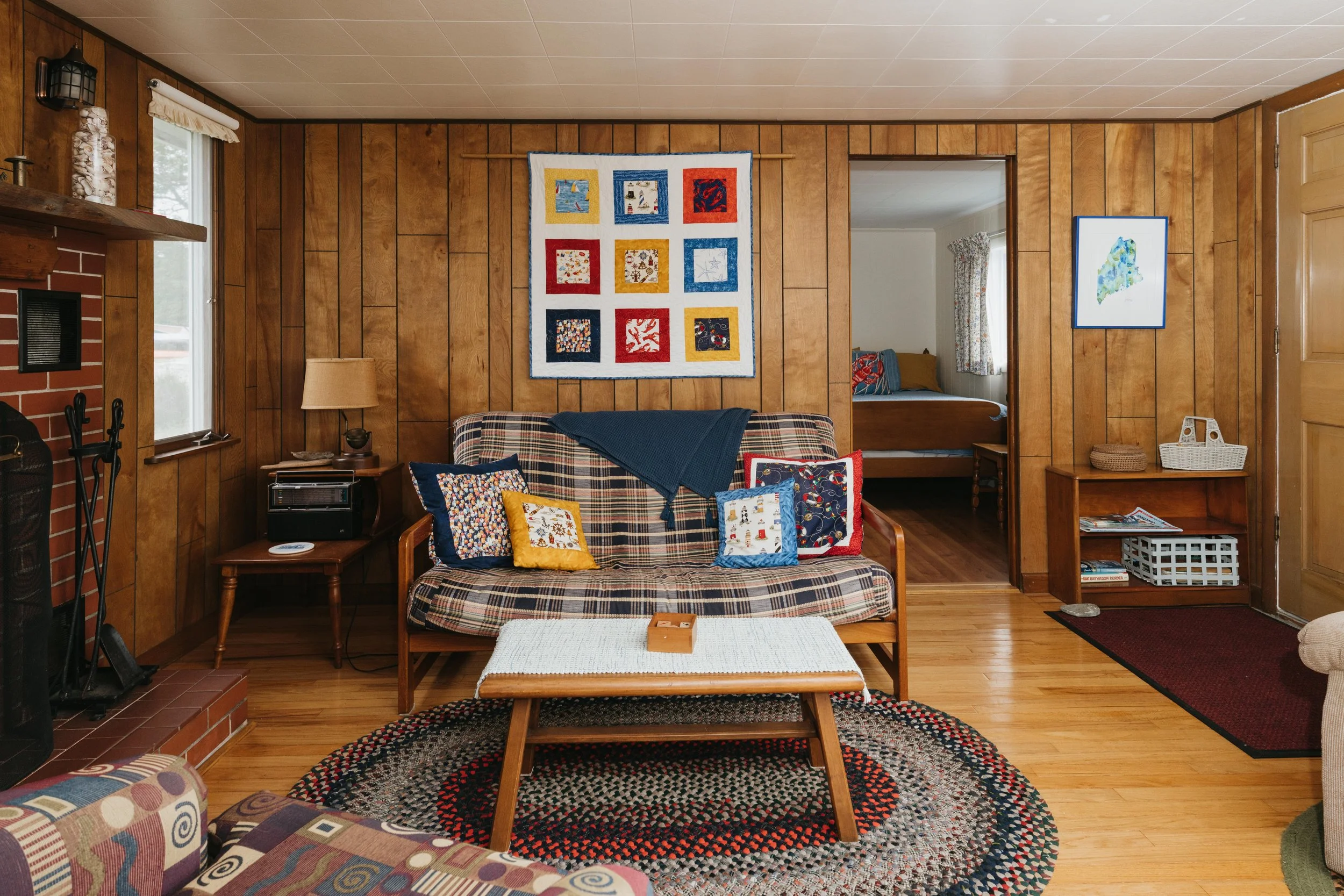 Living room with wood-paneled walls, fireplace, plaid sofa with multiple decorative pillows, wooden coffee table, rug, and artwork on the walls, with view into a bedroom.