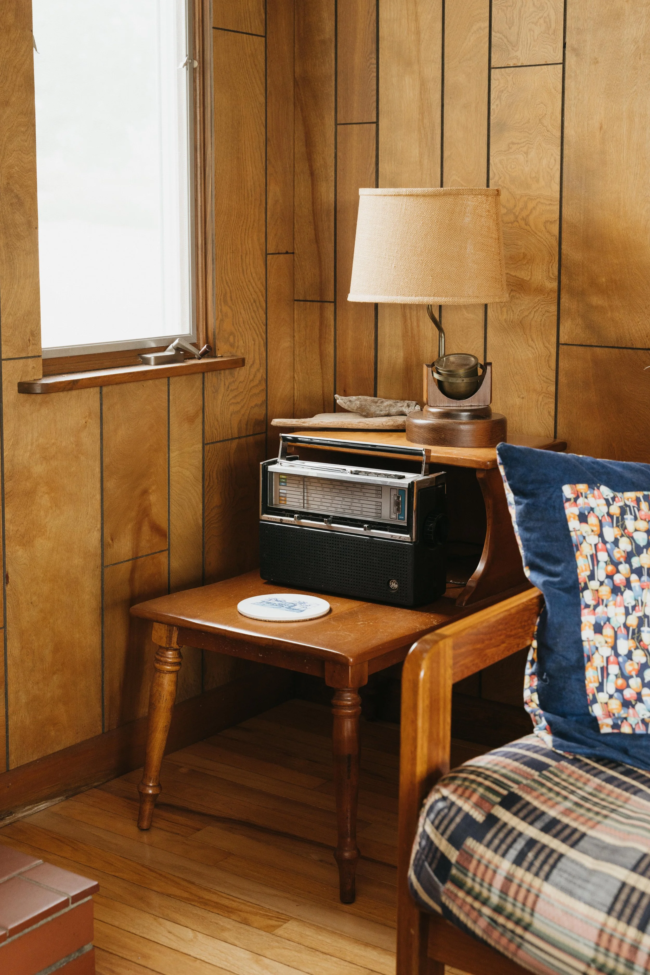 A wooden side table with a vintage radio, a white ceramic dish, and a table lamp with a beige lampshade on top. There is a window with frosted glass and wood trim in the background. Part of a cushioned wooden chair with a patterned pillow is visible 