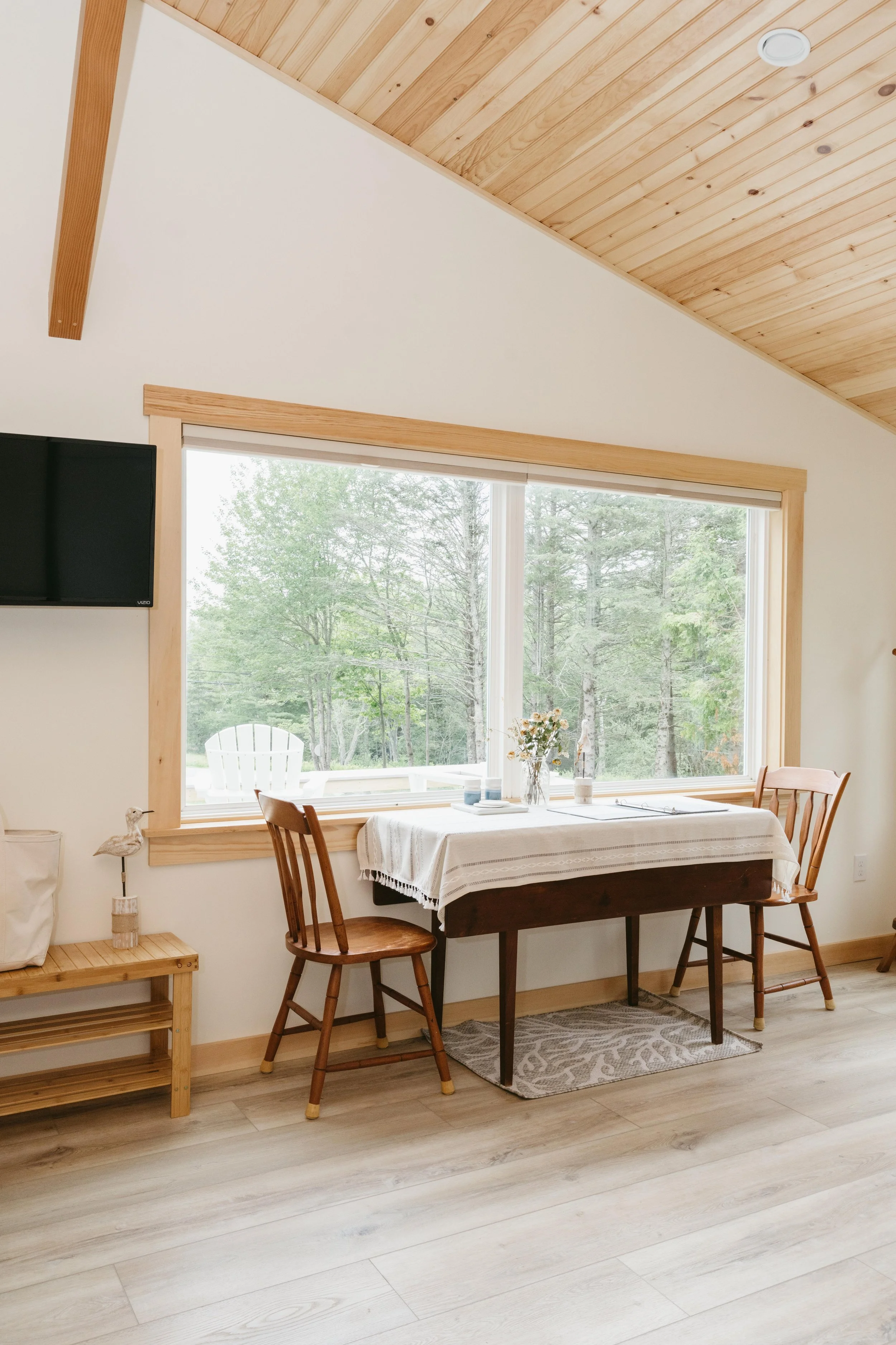 Room with wooden ceiling, large window showing trees, table with tablecloth and two chairs, small shelf with decor, and television
