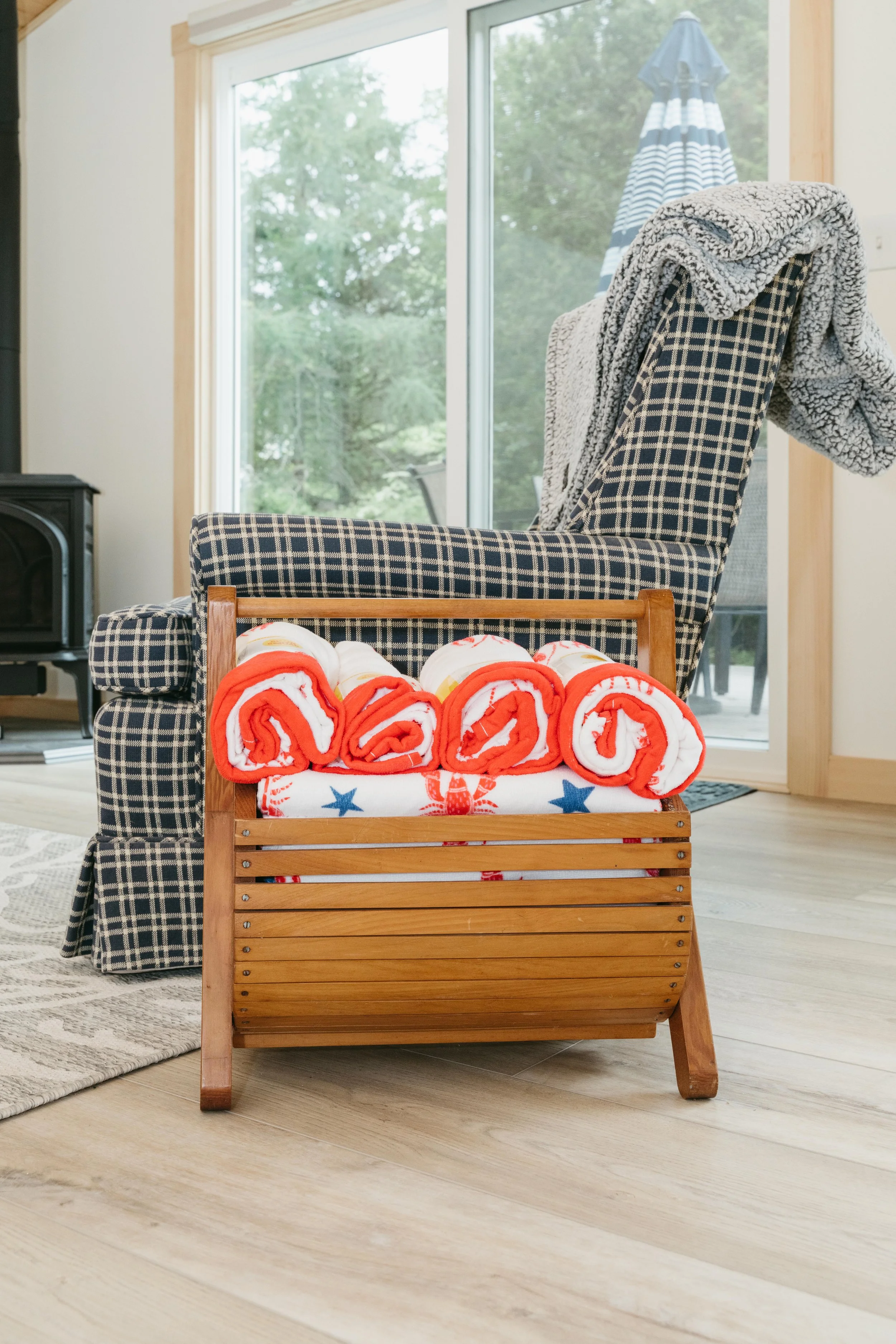 A wooden towel rack with red, white, and blue towels, placed in front of a checkered armchair with a gray fleece blanket draped over it, and a sliding glass door leading to an outdoor patio.