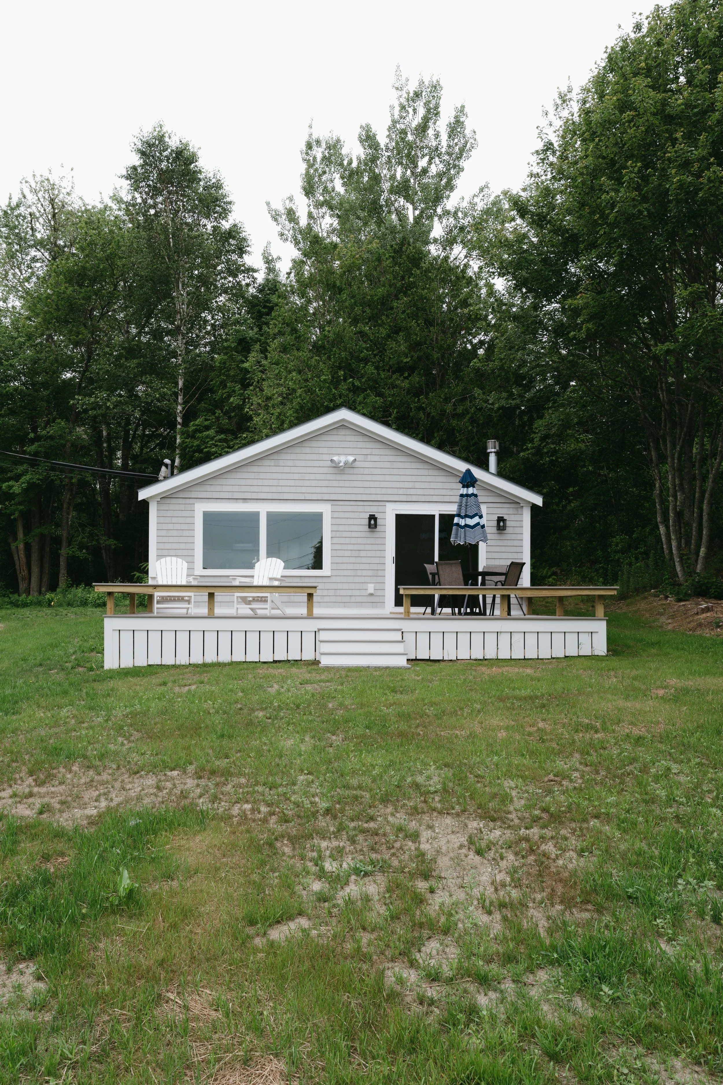 A small house with a porch, two Adirondack chairs, and a dining table with chairs and an umbrella, set against a backdrop of green trees.