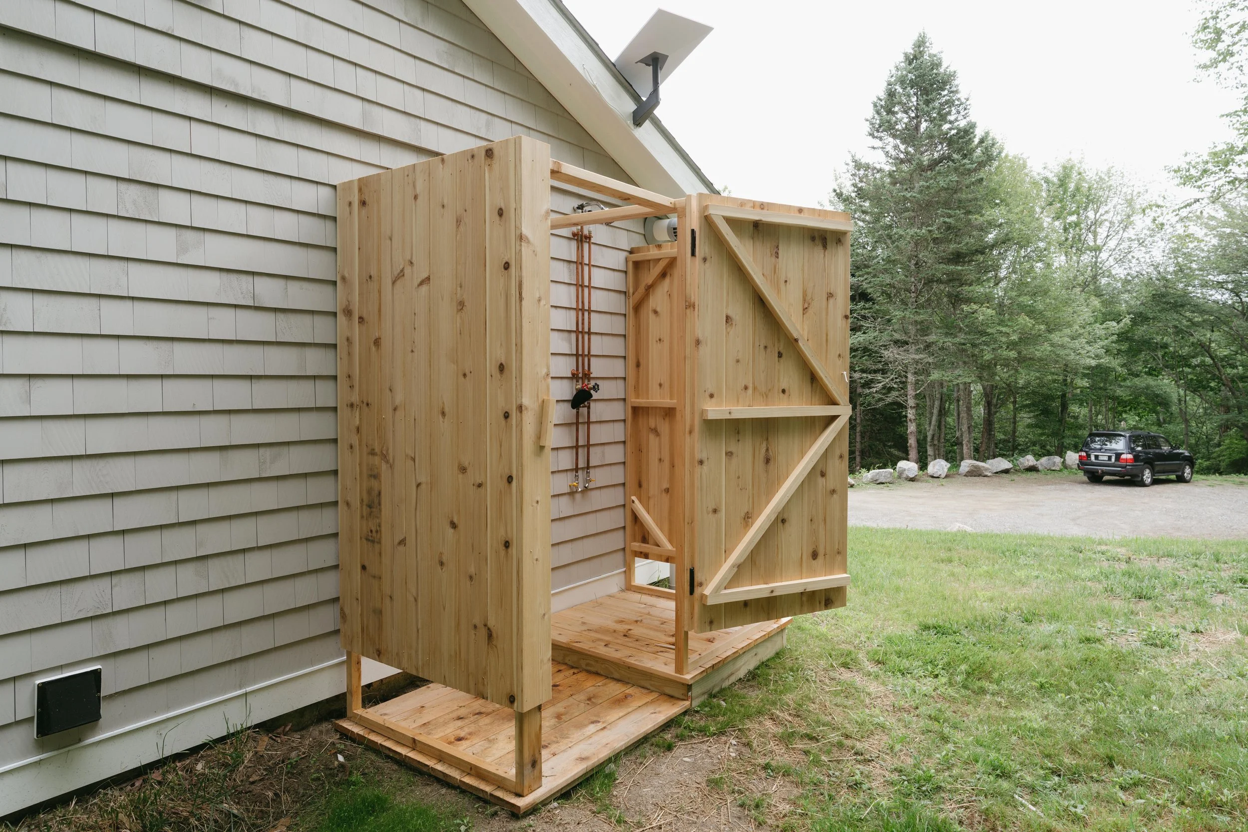 An outdoor wooden shower structure attached to a house with a large open door, overlooking a grassy yard and a parking area with a black SUV, surrounded by trees.