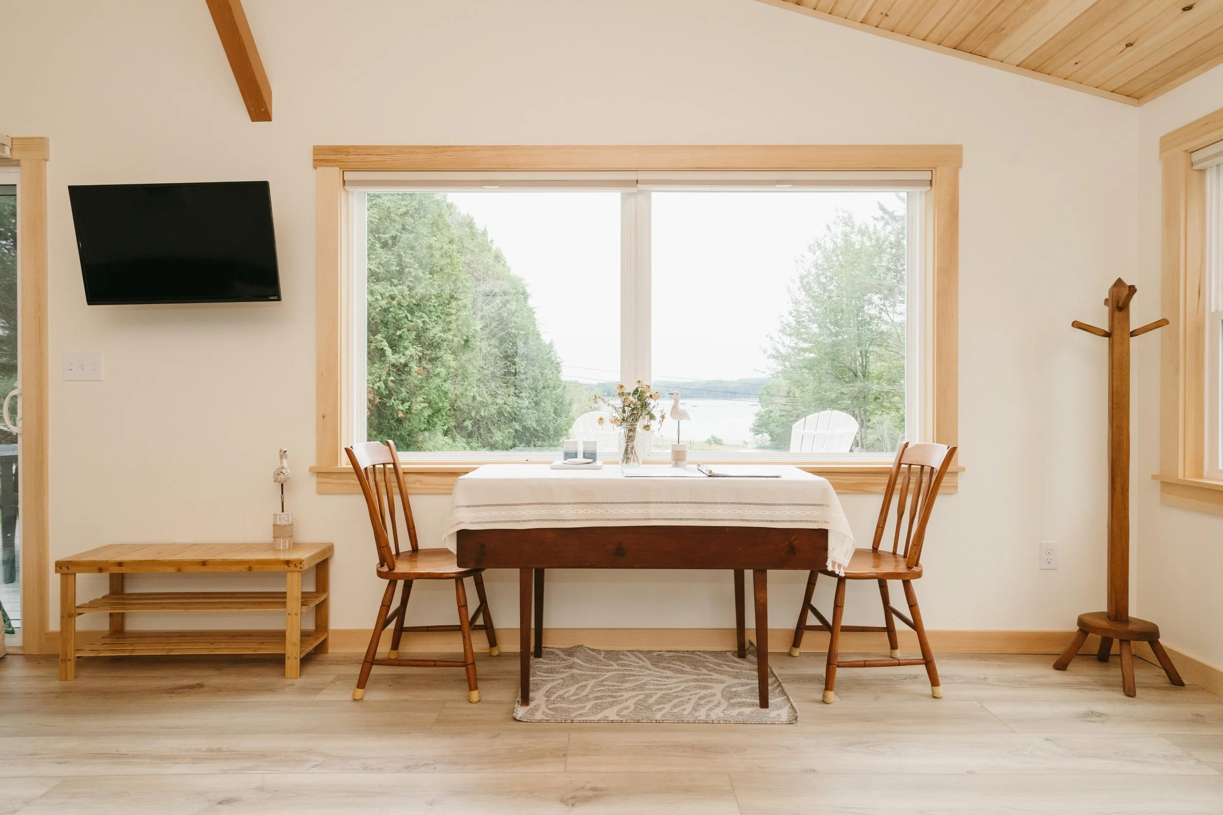 Dining area with a wooden table covered with a white tablecloth, flanked by two wooden chairs, in front of large window overlooking a green landscape with trees and water.
