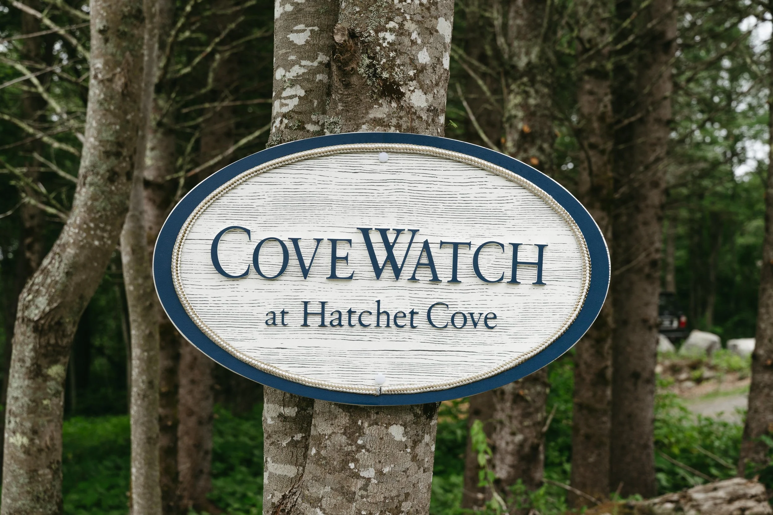 A sign that reads 'CoveWatch at Hatchet Cove' hanging on a tree trunk in a wooded area.