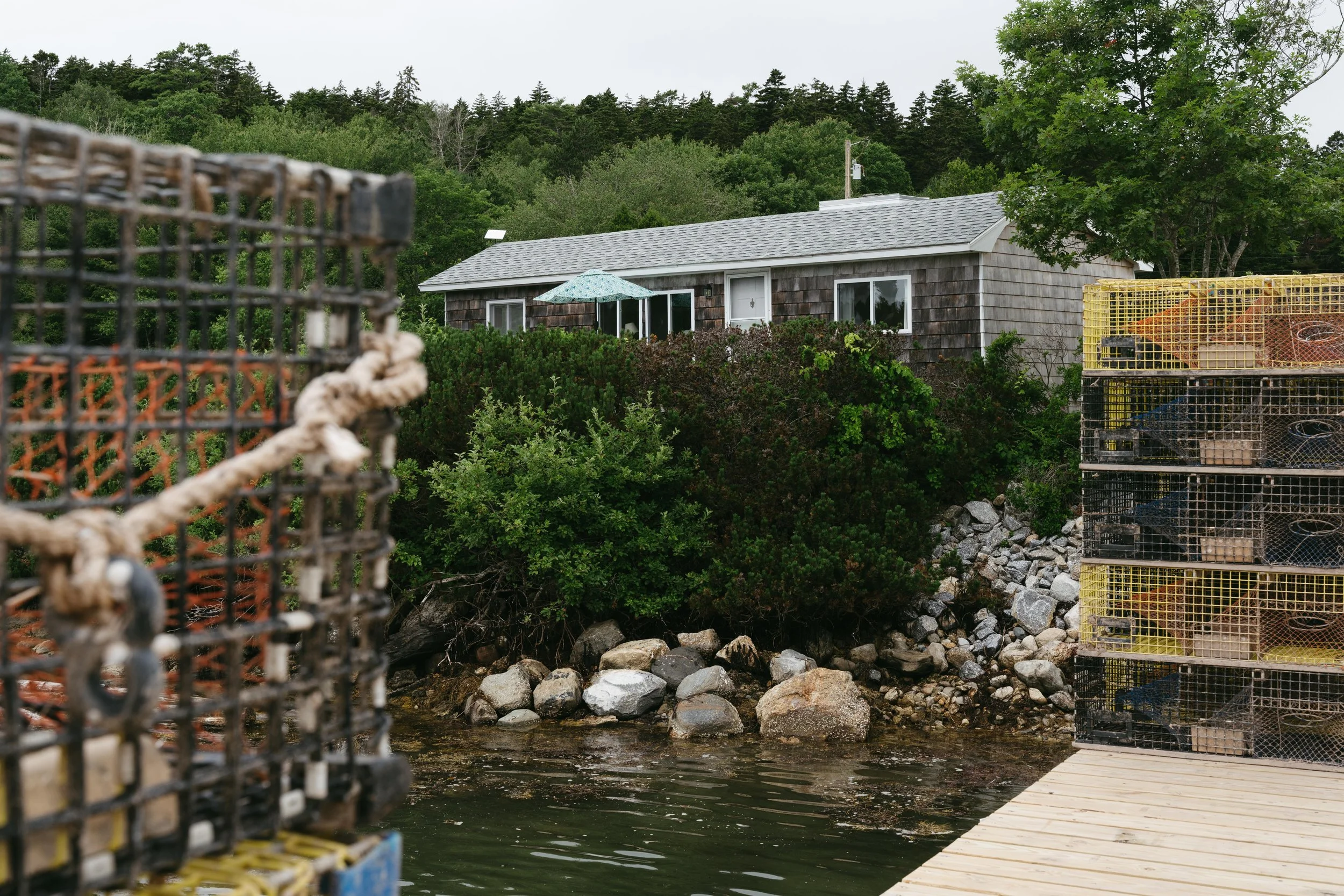 Lobster traps and crates near a waterfront with a house and trees in the background.