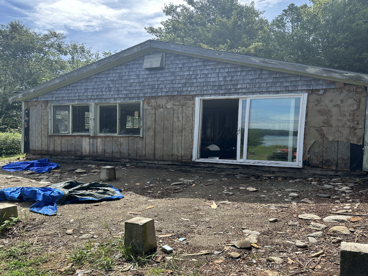 A house under renovation with exposed wood siding and a sliding glass door, situated on uneven dirt ground with scattered rocks and construction debris, near a body of water and surrounded by trees.