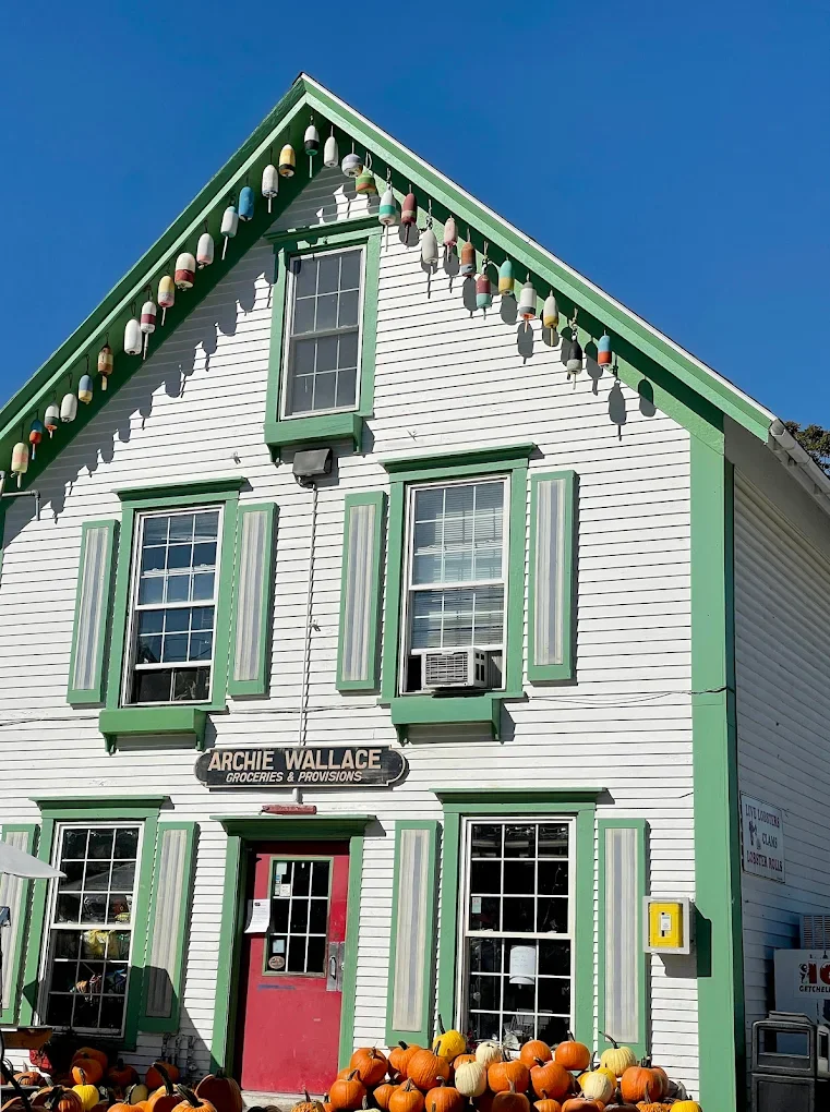 White wooden building with green trim, decorated with colorful string lights, pumpkins in front, and a sign that reads 'Archie Wallace Groceries & Provisions'.